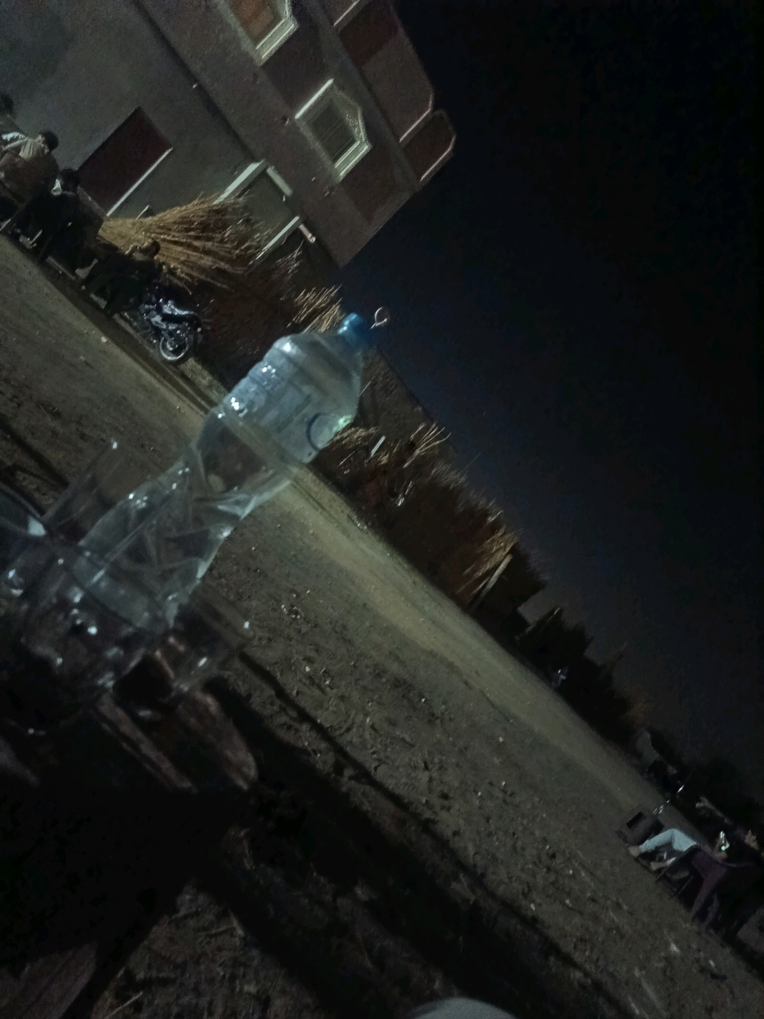 The image shows a dimly lit, outdoor scene at night, possibly in the city of Bani Mansour, Egypt. In the foreground, a partially empty plastic water bottle is prominently featured, lying on its side on the ground. The bottle appears to be clear with a blue cap. The ground is a mixture of dirt and gravel, with some scattered debris. In the middle ground, a motorcycle is parked, and a large pile of straw or dried vegetation is visible. A building with illuminated windows is present in the background, suggesting it is a residential or commercial structure. Several individuals are dimly visible in the background, some standing and others appearing to be seated or lying down. One person is clearly visible lying on the ground in the far right, with their legs extended and wearing light-colored pants and a dark top. The overall impression is a casual, informal gathering outdoors at night.