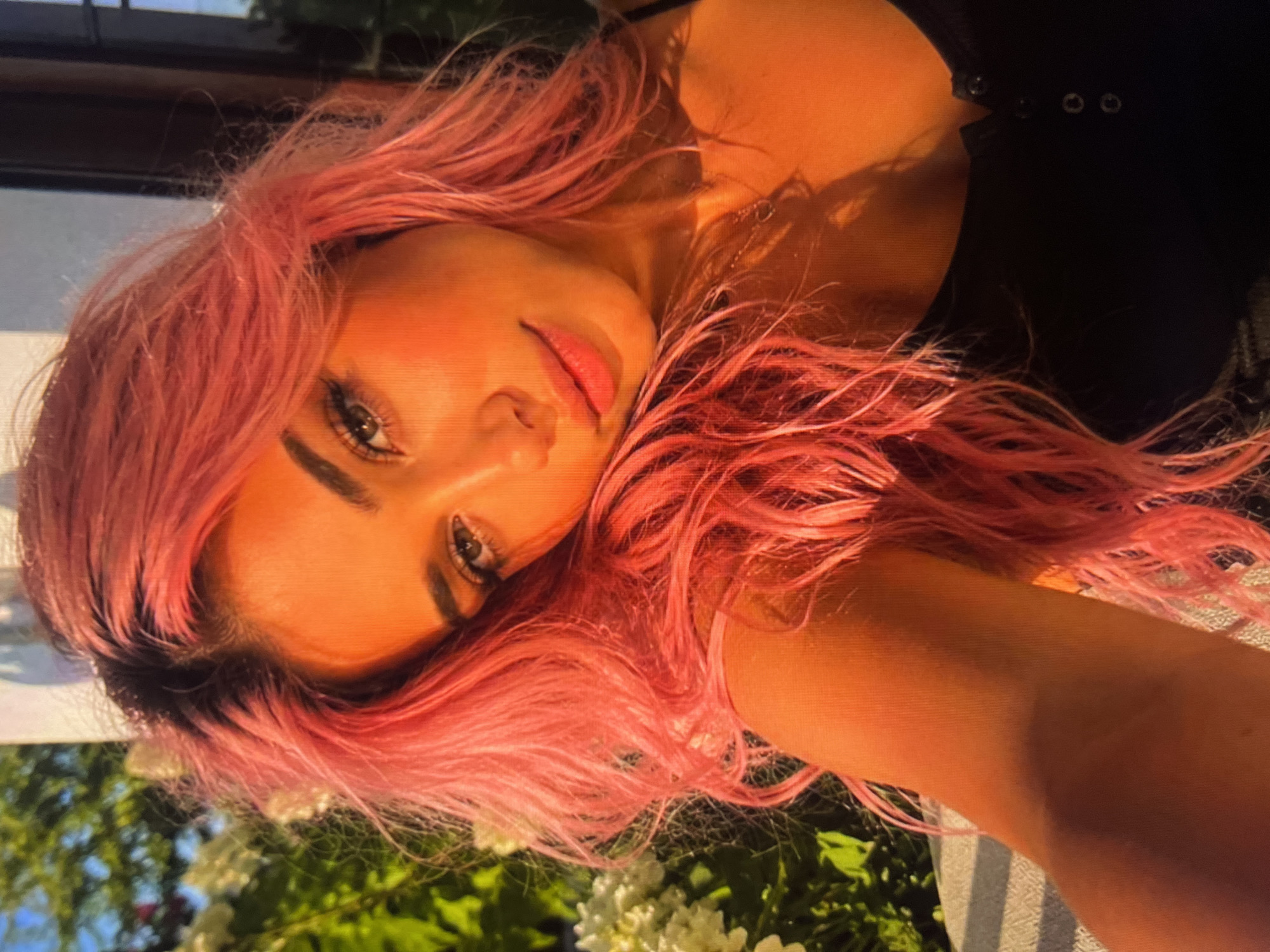 An outdoor portrait shows a young woman with bright pink hair, looking towards the camera. She is wearing a black, buttoned top. The lighting suggests it is either sunrise or sunset, casting a warm, orange glow on her face and skin. Behind her, out of focus, are green foliage and white flowers. The background is blurred, indicating a shallow depth of field.