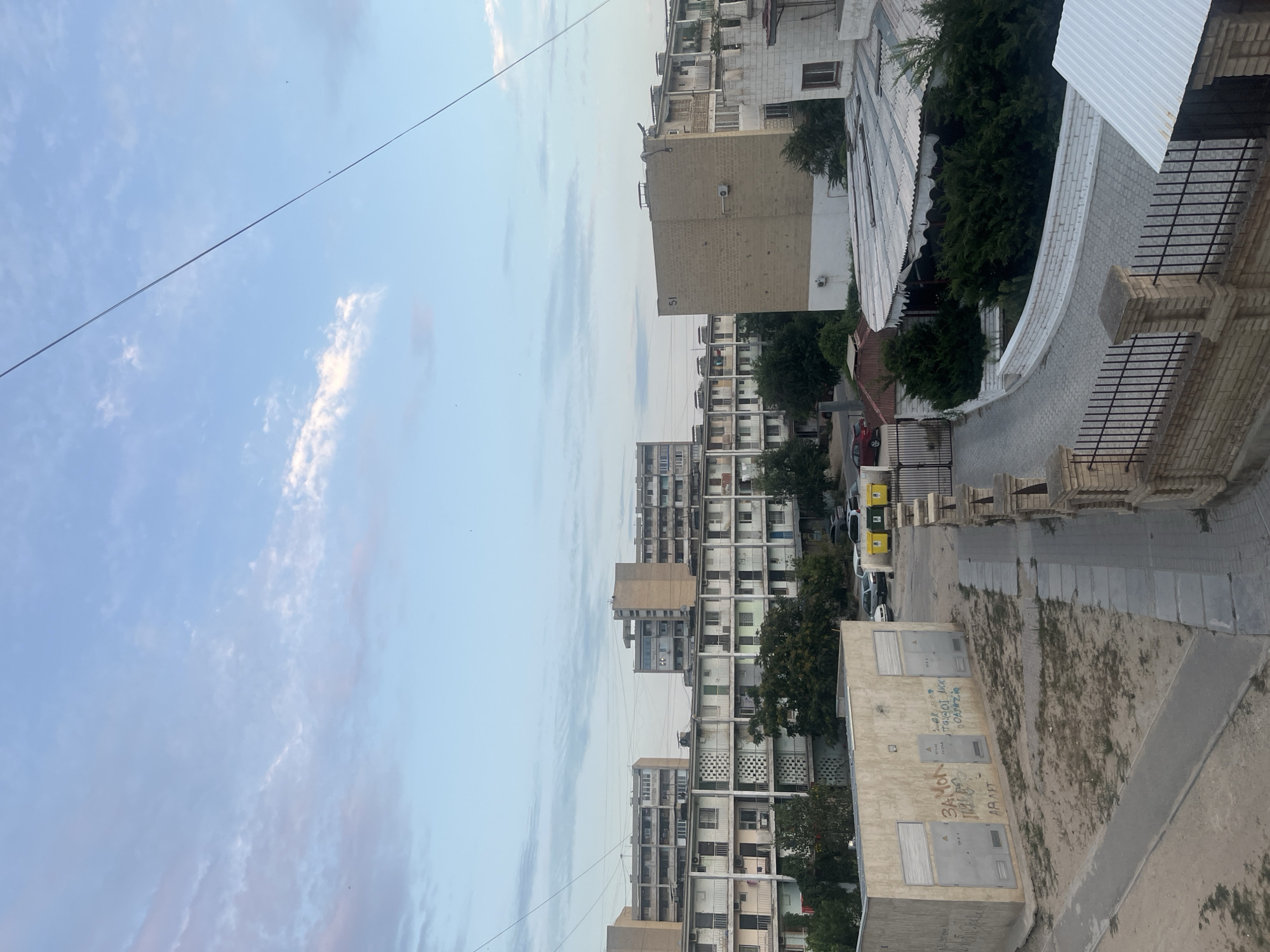 This image captures an outdoor scene in Aktau, Kazakhstan, likely during the early evening due to the soft lighting and pale sky with wispy clouds. The view is dominated by multi-story apartment buildings with rows of balconies, characteristic of Soviet-era architecture. These buildings are constructed from concrete and appear to be aged, with some showing signs of wear and tear. In the foreground, a low, rectangular building with two large doors is covered in graffiti. Adjacent to it is a paved walkway leading to a street where several cars are parked. Further down the street, more apartment blocks stretch into the distance, some of them taller than others. To the right of the street, a building with a curved, corrugated roof is partially visible, surrounded by dense green bushes. A metal fence with brick pillars separates the foreground area from a paved driveway. The overall impression is of a residential area with an urban, somewhat utilitarian aesthetic. No people are visible in the frame.