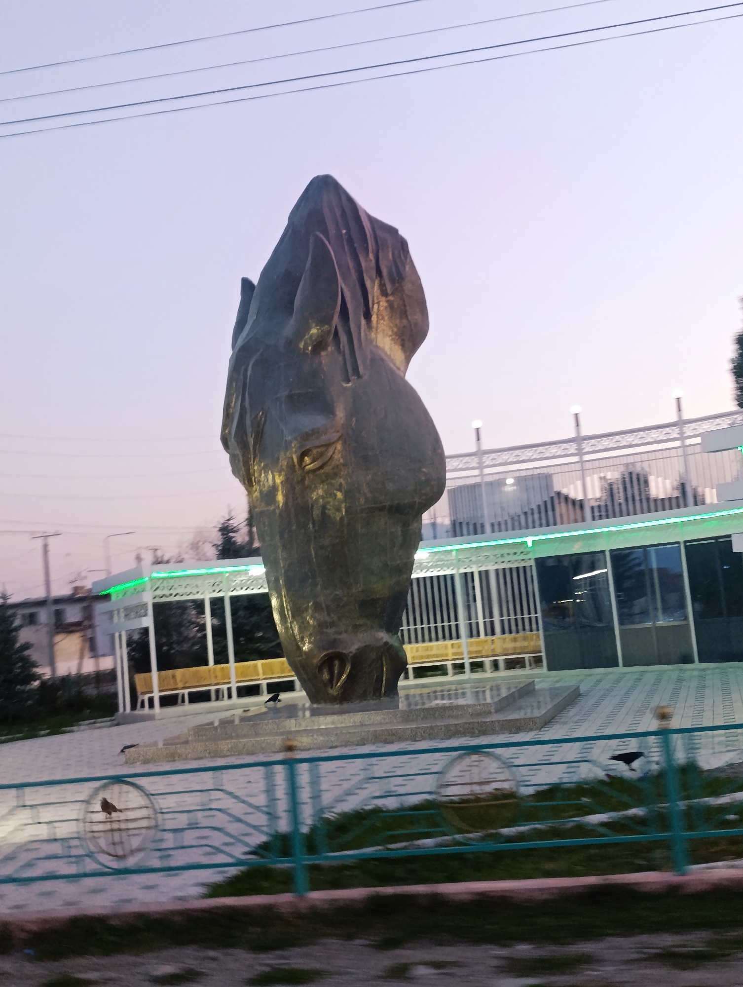 In the city of At-Bashy, Kyrgyzstan, a monumental sculpture of a horse's head dominates the foreground. The sculpture, made of a dark, metallic material, is positioned centrally and appears weathered. Behind the sculpture, a modern building complex is visible, featuring white latticework, glass windows, and green neon lighting. Benches are placed in front of the building, and a pathway leads up to it. A teal-colored metal fence separates the foreground from the pathway. Several birds are present in the scene: one small bird is perched on the fence, and two dark birds are on the ground near the sculpture's base. The sky above is a pale, twilight gradient.