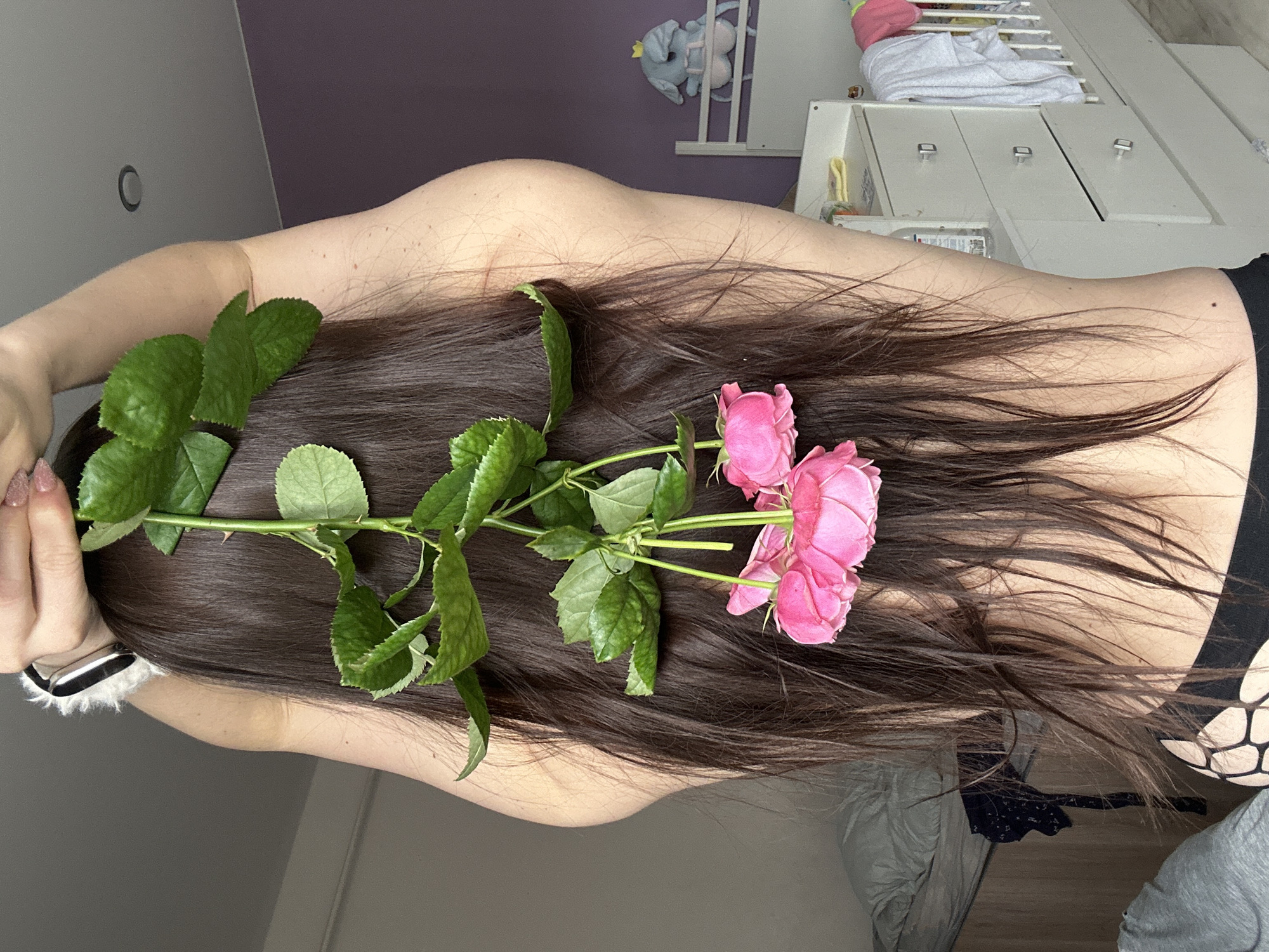 A woman is positioned with her back and head toward the camera, her long, brown hair fanned out. A stem of pink roses with green leaves is placed along the length of her hair. The woman is wearing a black garment with a pattern near her lower back. The background includes a purple wall, a white dresser with drawers, and a crib with white bedding. The location is not evident from the image, and there is no indication that it is in the city of Rusalevka, Russia.