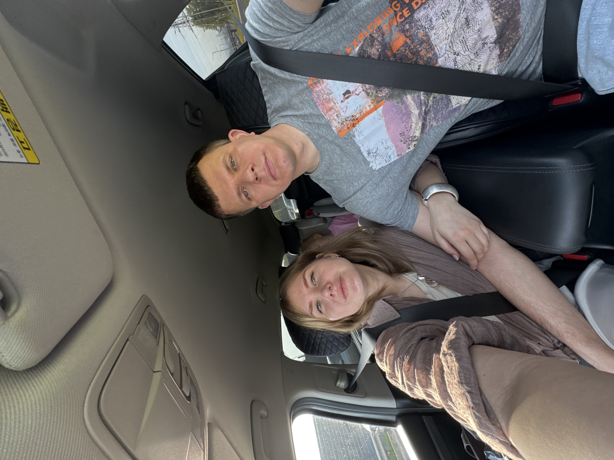 The image shows two individuals, a man and a woman, seated in a car. The man is in a gray t-shirt with a graphic design and is wearing a seatbelt. The woman is wearing a light brown top and is also wearing a seatbelt. The interior of the car is visible, including the dashboard, sun visor, and seats. The location cannot be determined as Lipaki, Russia, without additional context within the image.