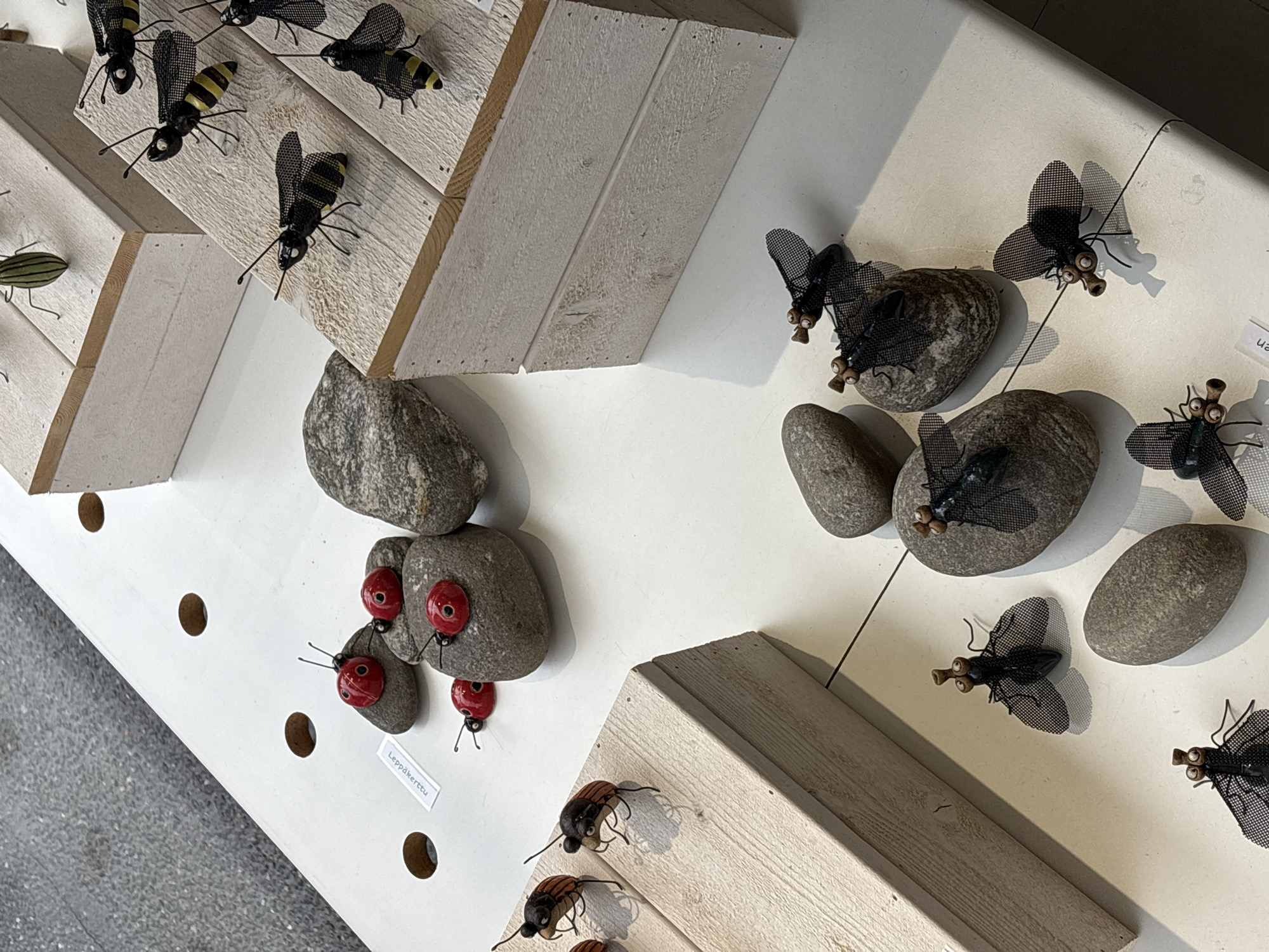 The image shows an outdoor market stall in Pori, Finland, displaying various handcrafted insect figures. The stall is set up on a white table with a textured surface. The figures are primarily made of metal and are designed to resemble ladybugs, bees, and flies.
In the foreground, several metal flies with mesh wings and protruding eyes are positioned on grey stones. To the left, two more flies are placed on a light-colored wooden display stand. Further left, on a similar wooden stand, are two metal beetle figures with orange and brown coloring.
In the midground, a cluster of red ladybug figures, made of what appears to be glass or polished stone, are perched on grey stones. A small white label with text in a foreign language is visible near these ladybugs. Behind them, on tiered wooden platforms, are several black and yellow striped bee figures. One bee is in the process of being placed on a platform by an unseen hand.
In the background, more wooden platforms hold additional insect figures, including a green and black striped insect. The overall setting suggests a vendor selling artisanal crafts at a public market. The lighting is bright, casting shadows on the table.