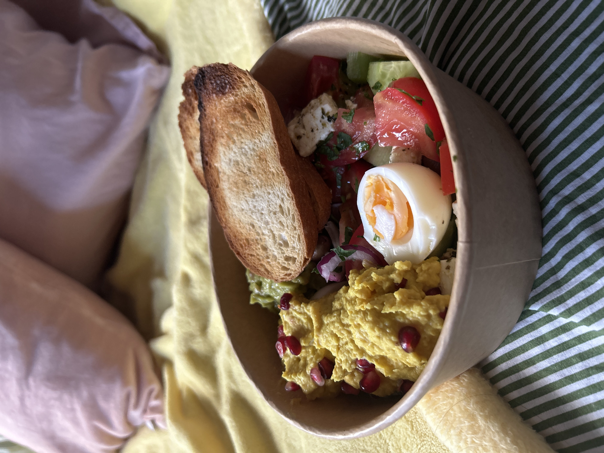 The image presents an overhead, slightly angled view of a meal contained within a disposable, light brown paper bowl. The scene is set indoors, within the city of Sofia, Bulgaria. The bowl rests on a fabric exhibiting parallel green and white stripes. To the lower left, a segment of a cream-colored, fuzzy, cylindrical object is partially visible. Behind the bowl, a textured, crinkled yellow fabric, likely a blanket or towel, covers the mid-ground. The upper background is occupied by a blurred, soft pink object, consistent with a pillow.
The meal within the bowl consists of several distinct components:
1. **Yellow Puree:** A significant portion of a vibrant yellow, pureed or mashed substance, possibly a lentil spread or turmeric-infused hummus, is situated on the left side, garnished with several dark red pomegranate arils.
2. **Green Spread:** Adjacent to the yellow puree, towards the top-middle, is a mass of green, chunky spread, consistent with guacamole or mashed avocado.
3. **Salad:** The right half of the bowl holds a mixed salad. This includes diced red tomatoes, uniformly cubed green cucumbers, white, crumbly cheese pieces (resembling feta) dusted with dark specks of herbs or pepper, thin slices of red onion, and chopped green herbs.
4. **Hard-Boiled Egg:** Positioned at the front-right of the bowl is a longitudinally bisected hard-boiled egg, displaying a solid white and a medium-cooked, orange-yellow yolk.
5. **Toast:** Two slices of toasted bread, characterized by golden-brown surfaces and darker, slightly charred edges, are placed on top of the food, leaning against the bowl's rim.