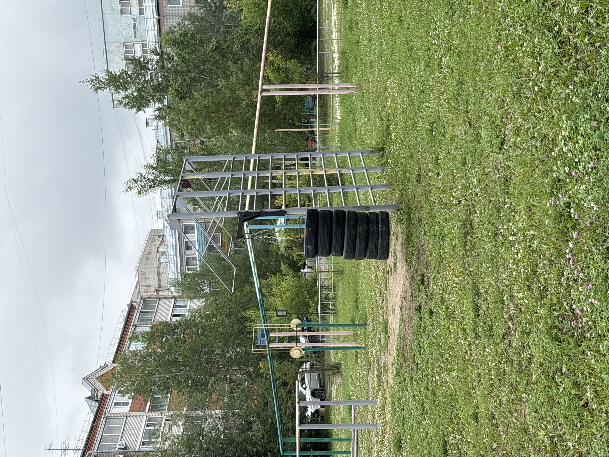 The image depicts an outdoor children's playground in Chigiri, Russia, with residential buildings and trees in the background. The playground features a metal climbing structure with a ladder, a horizontal bar, and a hanging tire punching bag. Several tires are stacked on the ground next to the punching bag. Green and pink metal bars form part of the playground equipment, along with a swing set. A white car is parked in the background. The ground is covered in grass and small white and pink flowers. The sky is overcast and gray.