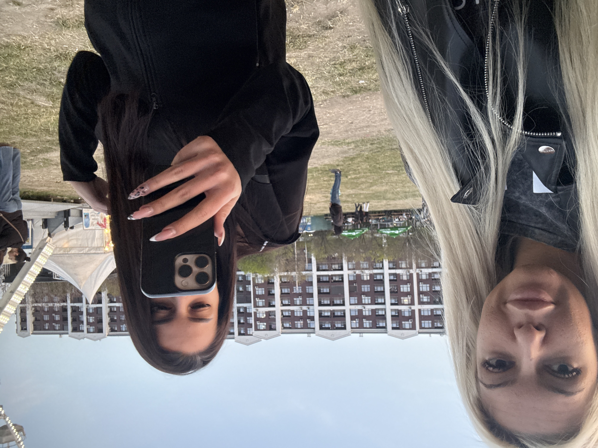 Two women are taking a selfie outdoors on a grassy area in front of a building with multiple stories and many windows. The woman on the left has dark hair and is holding a black phone up to take the picture. She is wearing a black jacket. The woman on the right has long blonde hair and is wearing a black leather jacket. In the background, there are tents, other people, and trees. The sky is visible in the background.
