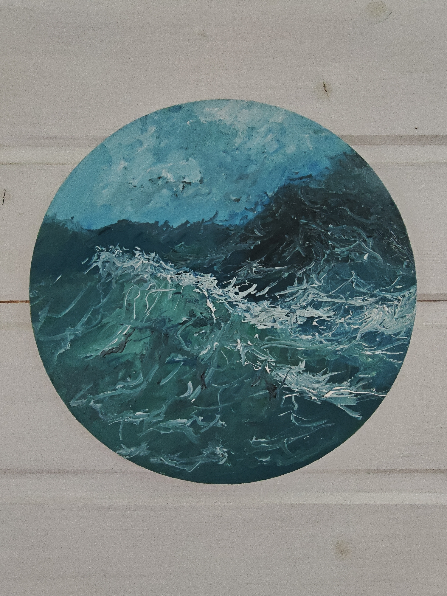 The image is a close-up, top-down shot of a circular painting of rough, turbulent ocean waves, rendered in shades of teal, deep blue, and white. The painting has a textured, impasto style, with thick brushstrokes creating a sense of movement and volume in the water. The waves appear to be breaking, with white foam cresting and swirling. The background of the painting shows darker, more abstract shapes that could represent a distant, shadowy coastline or the ocean depths. The painting is positioned on a light-colored, horizontal wooden surface, likely a wall or table, which shows visible grain and plank lines. There is no indication of the city of Koledino, Russia, or any other specific location within the image itself.