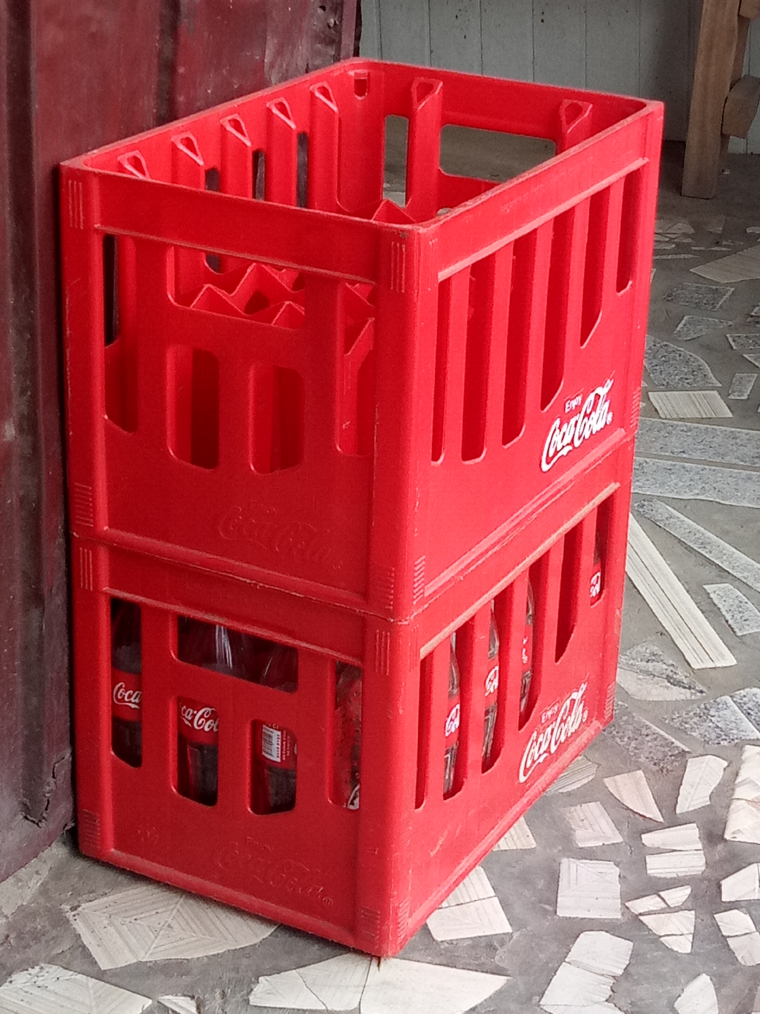 This media file shows two stacked red plastic Coca-Cola crates. The crates are full of Coca-Cola bottles. The top crate is empty, while the bottom crate contains bottles that are partially visible through the openings. The crates are positioned next to a dark red, textured surface, possibly a wall or a large object. The ground beneath the crates is a mosaic of light-colored stones and grey concrete.
Text visible on the crates includes "Enjoy Coca-Cola" and the Coca-Cola logo, which are printed in white. There are also some labels on the bottles in the lower crate, but the text is too small to be legible.
The scene appears to be outdoors, or in an open, covered area, given the ground material and the lighting. The lighting suggests it might be daytime, but there are no clear indicators of time of day or weather conditions. The overall impression is of a storage or display area, likely in a commercial or retail setting, given the branding. There are no people or animals in the image, nor is any specific activity taking place. The image is a still shot of the crates.