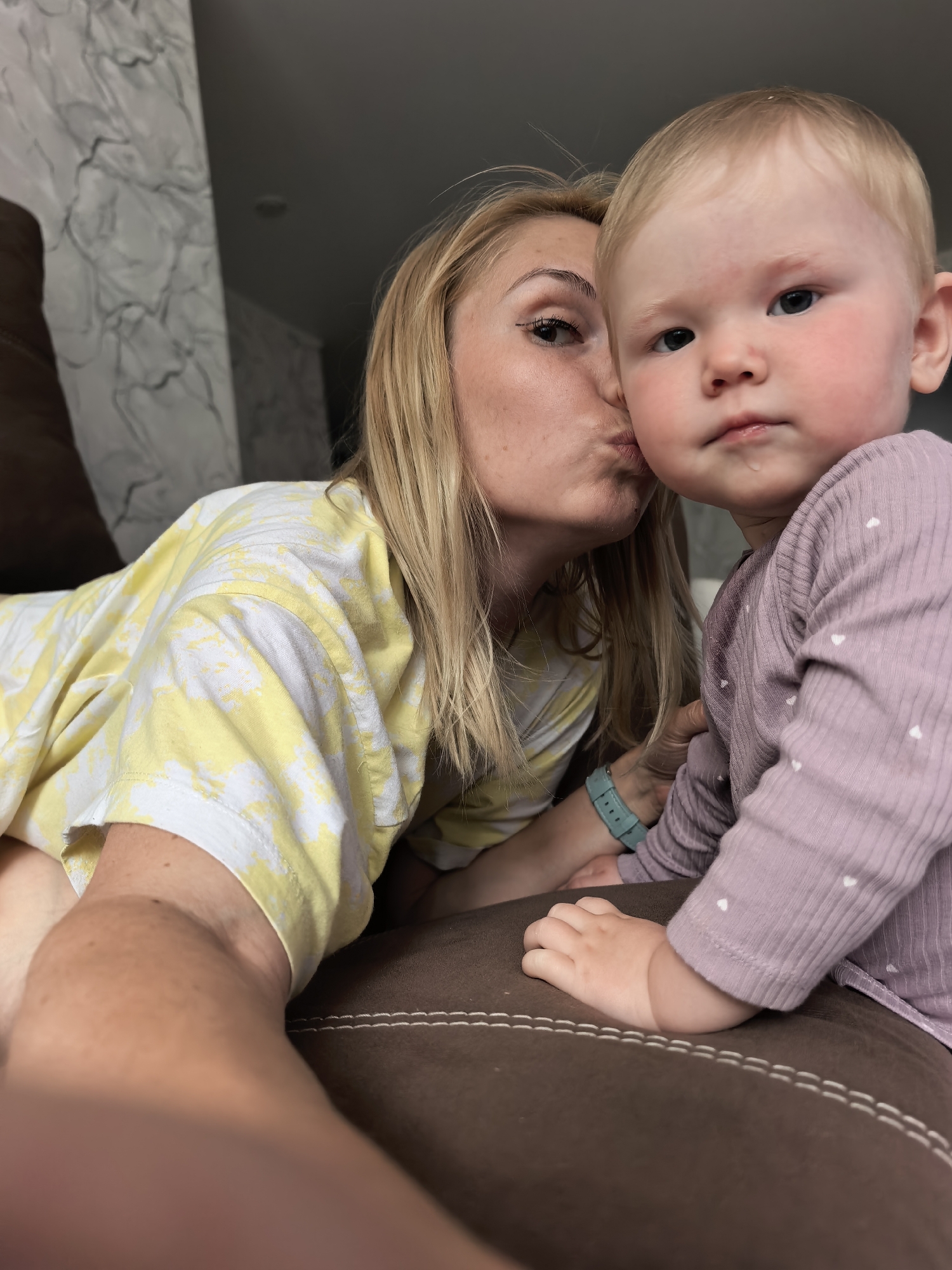 The image is a close-up, indoor selfie featuring an adult female and a baby. The adult female, positioned on the left, has blonde hair and is wearing a yellow and white tie-dye short-sleeved t-shirt. She is captured in the act of puckering her lips and kissing the baby's right cheek. Her visible left eye is made up with dark eyeliner. A light blue watch is visible on her left wrist.
The baby, appearing to be a toddler, is positioned centrally and to the right, looking directly towards the camera with a neutral expression. The baby has very short, light blonde hair, rosy cheeks, and an area of slight redness above the right eyebrow. A small droplet of liquid, likely saliva, is visible on the baby's lower lip/chin. The baby is wearing a light purple, long-sleeved, ribbed top adorned with small white heart patterns. The baby's right hand rests on a brown upholstered surface, which features white contrasting stitching along its edge, suggesting a couch or armchair.
In the background on the left, a wall with light-colored wallpaper is visible, featuring an abstract pattern of dark grey, wavy lines. The upper background to the right shows a plain grey surface, likely a ceiling or upper wall. The overall setting is a residential interior. There is no discernible visual information within the image itself to confirm a specific city or country, including Sterlitamak, Russia.