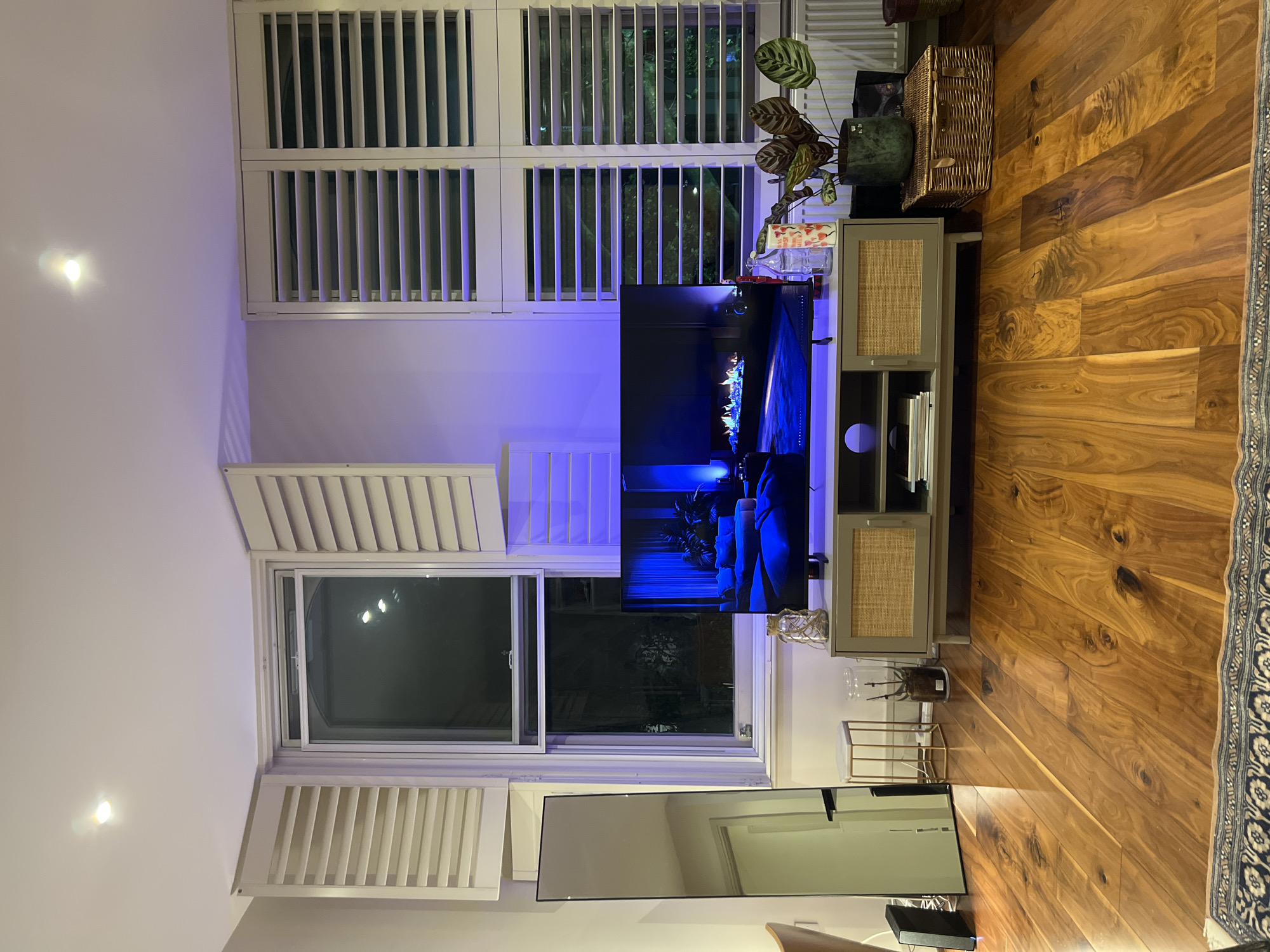 The image depicts a room with wooden flooring, a television set on a stand, and windows with white shutters. A plant sits atop the television stand. A mirror leans against the wall to the left of the TV. The lighting is a mix of natural light from the windows and an apparent blue light emanating from behind the television. There is no evidence to confirm or deny its location in London, United Kingdom. The scene shows no people or any specific actions or interactions.