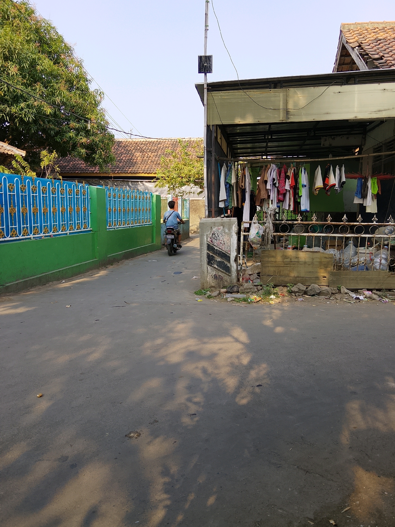 A man rides a motorcycle away from the camera down a paved road in Cirebon, Indonesia. The road is bordered on the left by a bright green fence with a decorative blue and gold railing. On the right, a structure with a metal roof overhang extends from a building. Clothes are hung on a line under the overhang, and various items, including plastic water bottles, are stored behind a decorative metal fence. The ground in front of this structure is littered with debris. Trees and houses with tiled roofs are visible in the background. The scene is illuminated by sunlight, casting shadows on the road.