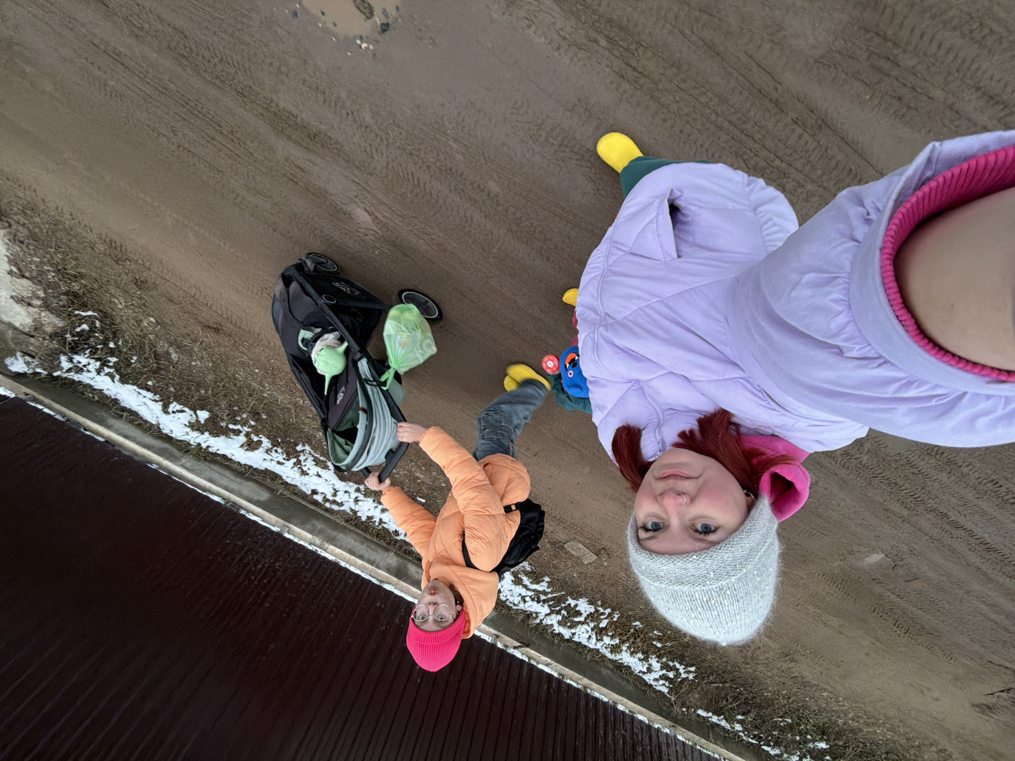 Two individuals stand on a muddy road in what appears to be a suburban area of Chaikovsky, Russia, as evidenced by the presence of snow, the architecture, and the type of road. The woman closer to the camera wears a light purple puffer jacket, a pink hooded sweater underneath, and a gray knitted hat. The other individual, presumably a young adult, wears an orange puffer jacket and a pink hat, and is pushing a black stroller. A child's legs and yellow boots are visible near the woman in the purple jacket. The ground is muddy and shows signs of vehicle traffic. Snow patches and some dry vegetation are present alongside the road. A dark-colored building or fence is at the bottom of the frame. The photograph appears to be taken from a high angle, potentially a selfie perspective.
