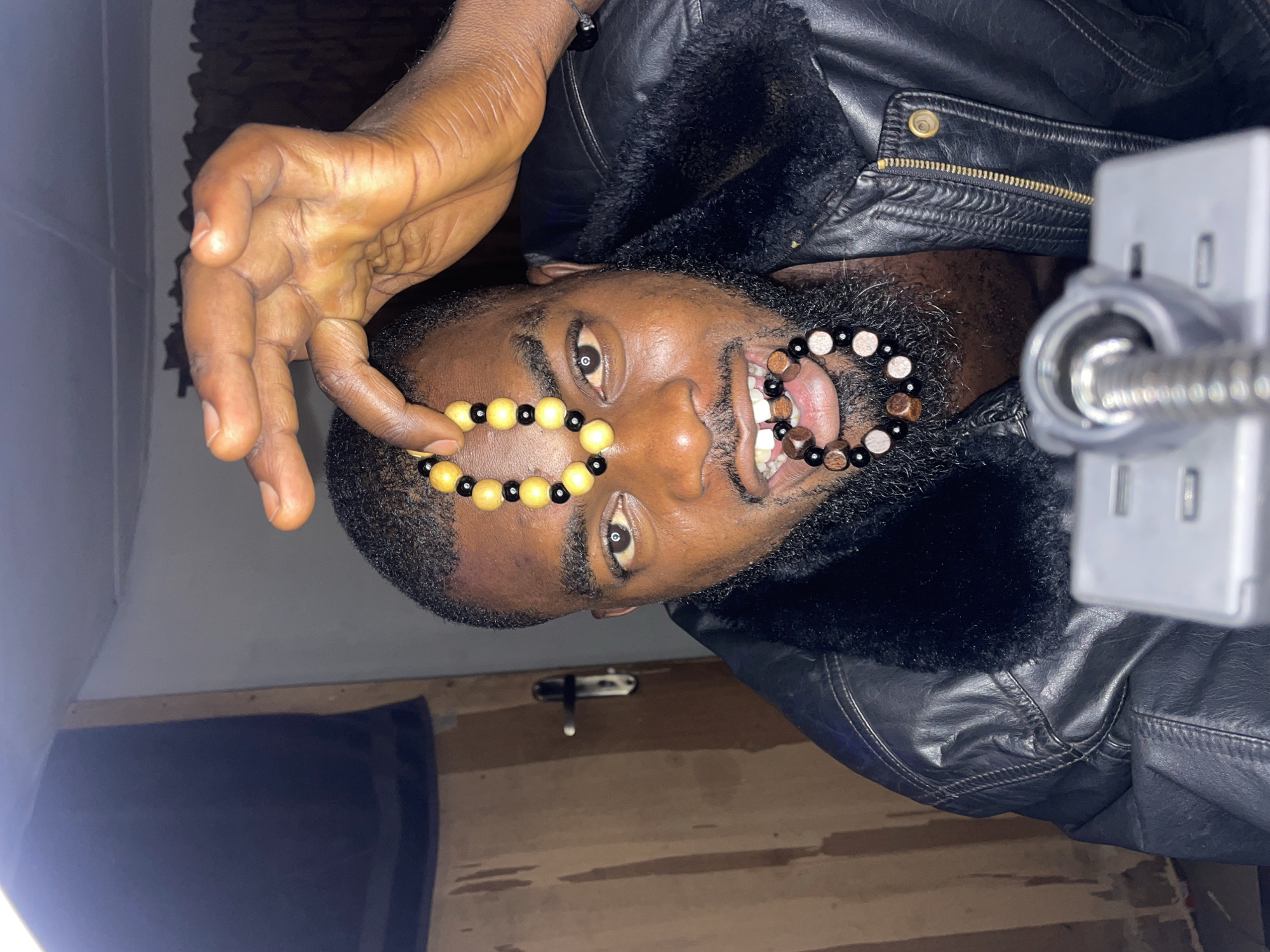 A Black man is shown in a vertically oriented photo, holding beaded bracelets in front of his eye and mouth as he looks at the camera. He wears a black leather jacket with a fur collar. The scene appears to be indoors. The location is Jos, Nigeria.