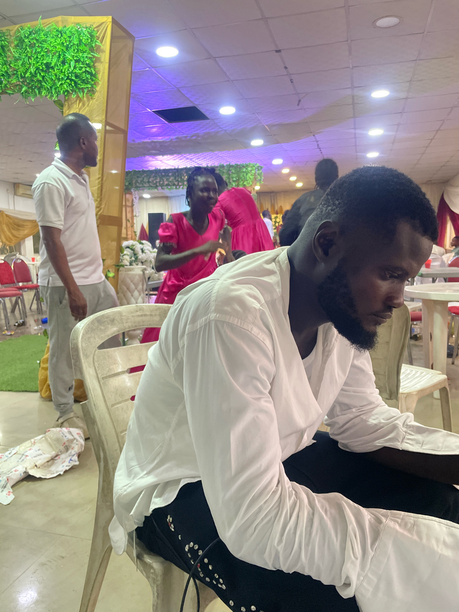 The image depicts an indoor gathering, likely a celebration or event, in Abuja, Nigeria. In the foreground, a Black male is seated, wearing a white collared shirt with the sleeves rolled up and black pants adorned with decorative studs. He has a beard and is looking down and to his right. Behind him, a Black female in a vibrant pink dress is partially visible, in motion. To the left, another Black male in a white polo shirt and grey sweatpants stands, facing away from the camera. The environment appears to be a decorated hall with green foliage accents, draped fabrics in gold and purple hues, and ceiling lights. Tables and chairs, some with red seats, are scattered throughout the space, suggesting a seating arrangement for guests. A stack of what appears to be food packaging or gift wrapping lies on the floor near the seated man. The lighting is a mix of overhead room lights and possibly colored ambient lighting.