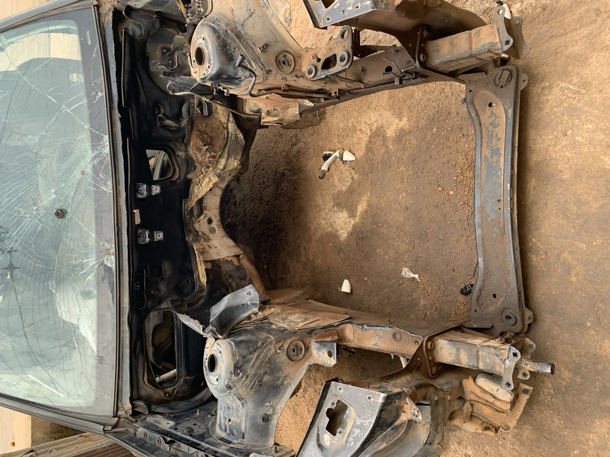 The image displays the extensively stripped and damaged frame of a vehicle, situated on reddish-brown dirt ground in Rafin Par, Nigeria. The vehicle's front section, encompassing the engine bay, is completely empty, with no engine or major components present. The bare chassis rails and front strut towers are visible, coated in significant amounts of reddish-brown dirt and exhibiting surface rust. The windshield, though still mounted within its frame, is severely shattered across its entire surface with a dense pattern of cracks. Sections of deteriorated insulation material are visible hanging from the firewall and inner fender areas. Scattered across the dirt ground around the vehicle frame are small pieces of white plastic and other unidentified debris. A light-colored building wall is partially visible in the upper left background. No individuals or ongoing activities are present within the scene.