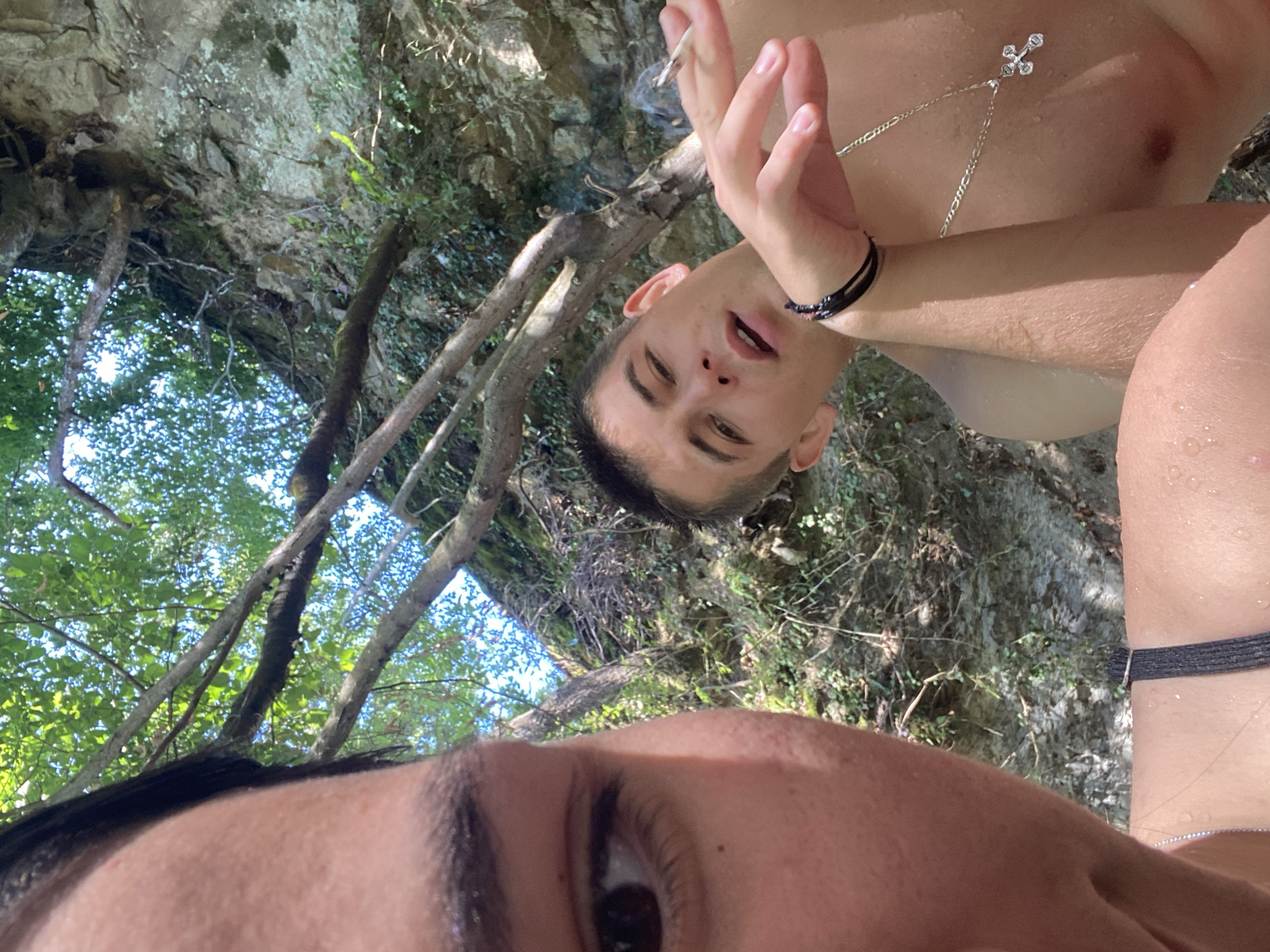 The image shows a low-angle, outdoor shot of two individuals near a rocky cliff face with dense foliage overhead, suggesting a natural, possibly forested, environment. The overall scene is suggestive of a recreational spot, perhaps near a body of water, given the wet appearance of the skin. The sky is visible through the leaves, indicating it's daytime.
The foreground features a close-up of a person's face and shoulder, partially obscuring the view. Their eye is visible, looking to the left. The skin appears wet, and they are wearing a black strap, possibly part of a bathing suit.
In the midground, another person is visible, also shirtless and with wet skin. They are holding a cigarette to their lips, with smoke rising. This individual is wearing a black bracelet on their left wrist and a silver chain necklace with a cross pendant. Their gaze is directed slightly upwards and to the left, towards the person in the foreground.
The background consists of a rough, textured rock face covered in patches of green vegetation and ivy. Several tree branches, some bare and others with leaves, extend across the frame, casting shadows and creating a dappled light effect. The overall impression is one of a secluded, natural setting, possibly a riverbank or a cliffside area.
While the image itself does not contain explicit visual cues to confirm the location as Debnevo, Bulgaria, the provided context states this setting. Without further information or visual landmarks, it's impossible to definitively identify Debnevo from the image alone. The scene depicts a candid moment, possibly a selfie, capturing two people enjoying an outdoor activity in a rugged, natural environment.