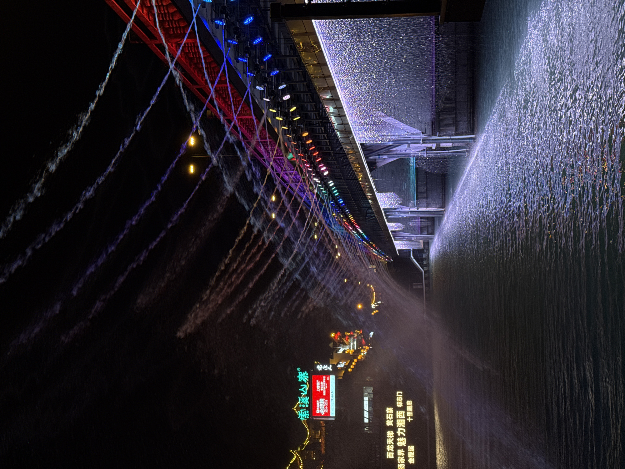 The image shows a nighttime view of a bridge in Wulingyuan, China, decorated with a colorful array of lights that transitions from red to blue along its length. Water cascades from the bridge into the dark water beneath, creating a dramatic effect. The water streams are illuminated by the colored lights, giving them a streaked appearance. On the shore, there are lit-up signs and buildings, including a large sign with Chinese characters. The area appears to be a tourist attraction with a scenic backdrop.