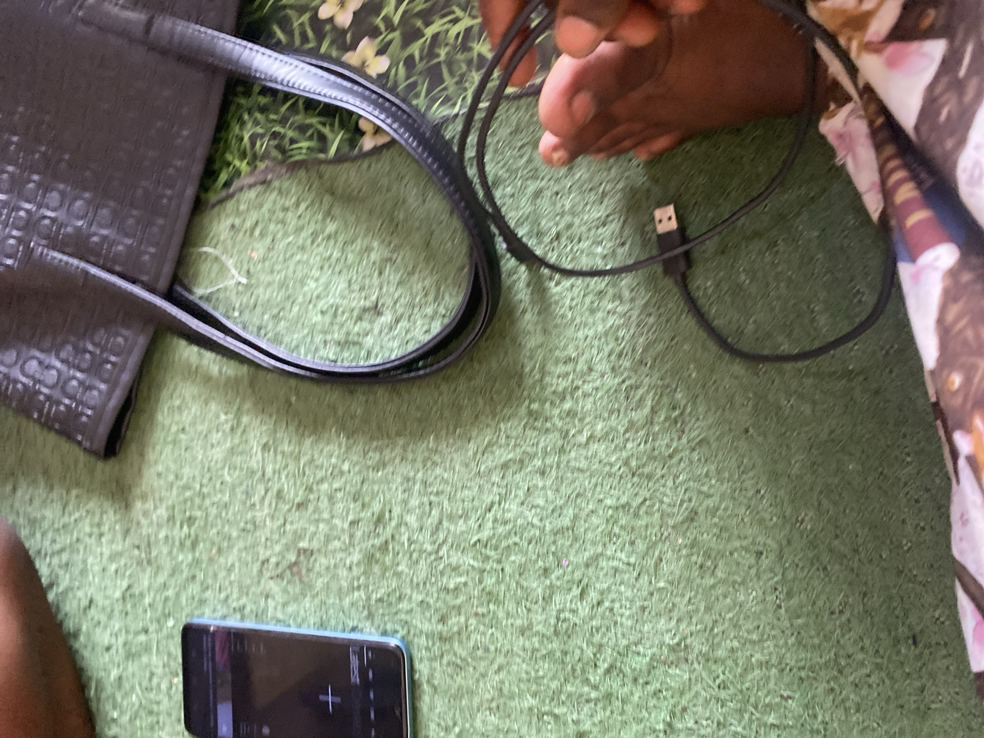 An overhead shot shows a black tote bag, a cellphone, and a USB charging cable on a green carpet. The tote bag, which has a textured pattern, is on the upper right side of the frame. A light blue cellphone with a dark screen is on the left. The screen displays a white plus sign. A black USB cable, with a silver connector on one end, is on the right side of the frame, in front of a person's foot. The carpet is short and appears to be artificial grass. This image was taken indoors.