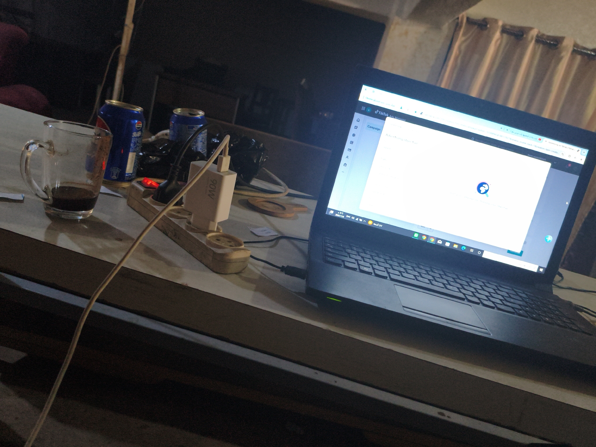 A laptop is open on a desk, displaying a webpage with a circular loading icon. Next to it is a glass mug with dark liquid, two cans of Pepsi, and a power strip with a white adapter labeled "M06" and a red indicator light. The scene is dimly lit, suggesting an indoor setting possibly in Gaza, Palestinian Territories, with a curtain visible in the background.