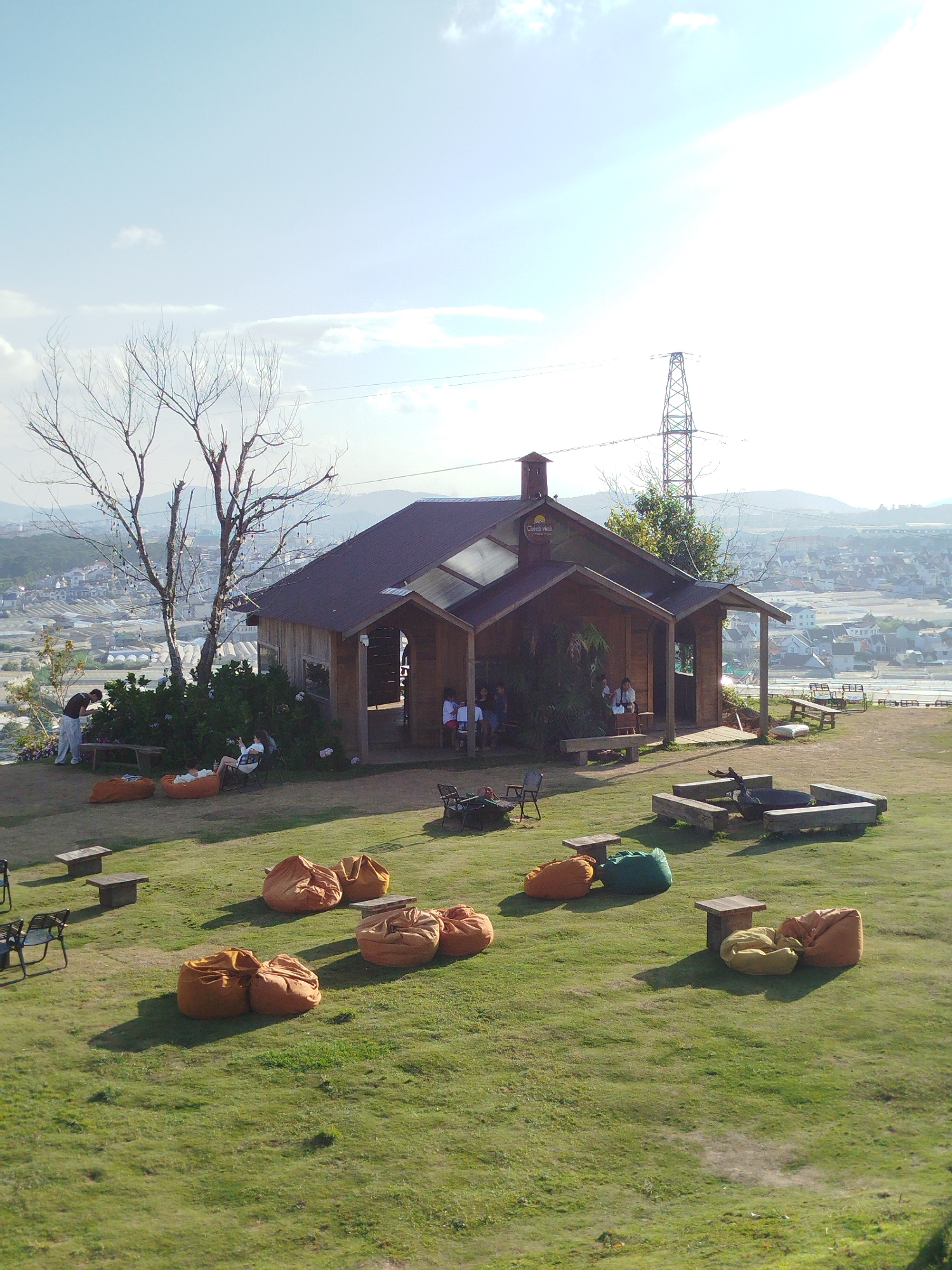 The image depicts a picturesque outdoor seating area at a café or restaurant, likely located in Lam Vien - Da Lat, Vietnam, given the characteristic hilly landscape and architecture. The scene is set on a grassy hill overlooking a sprawling town or city with numerous buildings, some appearing to be greenhouses. Dominating the foreground are numerous beanbag chairs in various shades of orange, yellow, and green, scattered across the lawn. A few small wooden tables are placed among the beanbags. In the mid-ground, a rustic wooden building with a brown roof serves as the main structure, possibly a café. Several people are visible sitting on chairs and beanbags outside the building, engaged in conversation or relaxation. A large, bare tree with decorative lights strung between its branches stands to the left of the building. Further back, a metal electrical pylon stands tall against the bright, hazy sky, a common sight in the region. The overall impression is one of a serene and inviting outdoor recreational space with a scenic backdrop.