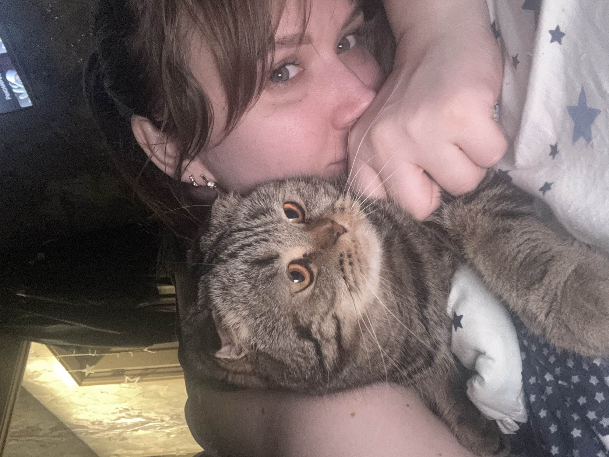 A woman with brown hair is holding a brown tabby cat in her arms. The woman has fair skin, grey eyes, and is wearing earrings. Her face is close to the cat's head. The cat has amber-colored eyes and is facing the camera. Both appear to be lying down. The background shows a room with a patterned white and blue fabric, possibly a blanket or bedding. Part of a dark object that appears to be a television is in the upper left corner of the photo. The location is not discernible from the image. The city of Chaikovsky, Russia, cannot be identified from the contents of this image.