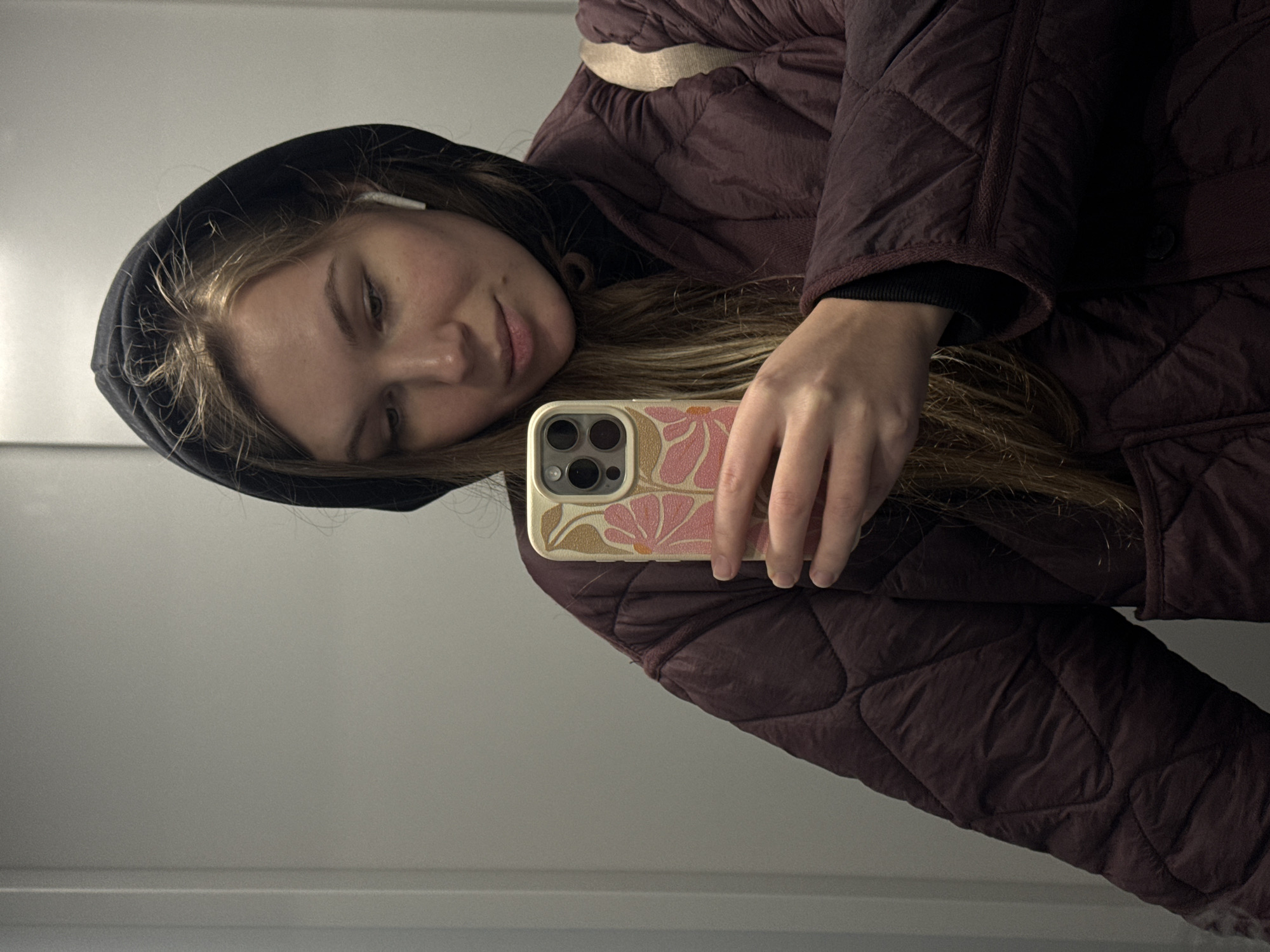 A young woman wearing a maroon quilted jacket with a black hood is taking a selfie. She is holding an iPhone with a floral patterned case in her right hand. An earbud is visible in her left ear. The background appears to be a grey wall. It's not possible to confirm the location as Vozdovac Urban Municipality, Serbia based on this image alone.
