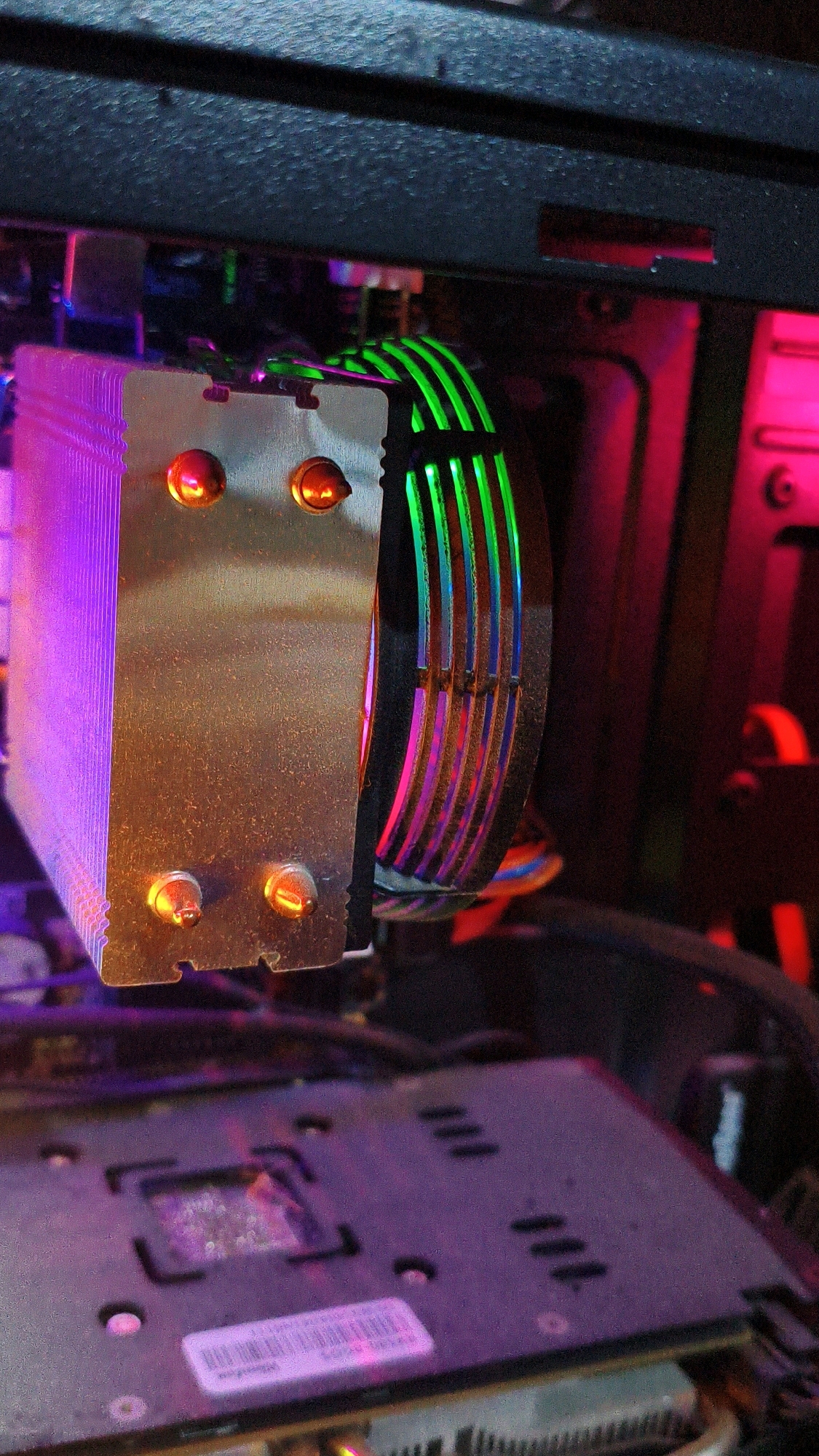 The image shows a close-up view of computer components within a computer case. A large metal heatsink with visible fins is positioned vertically, with two copper heat pipes extending from its base. Attached to the heatsink is a fan with RGB lighting that displays a gradient of green, blue, and pink hues. The fan blades are visible, along with wires connected to it. In the foreground, a portion of a motherboard is visible, showing the CPU socket area with some components and a sticker. The scene is illuminated by the RGB lighting from the fan and possibly other internal lights, casting purple and pink glows on the components. The background is dark and shows the interior structure of the computer case. There are no people present in the image, and no identifiable urban or natural environmental features are visible. The image is focused on the internal hardware of a computer.