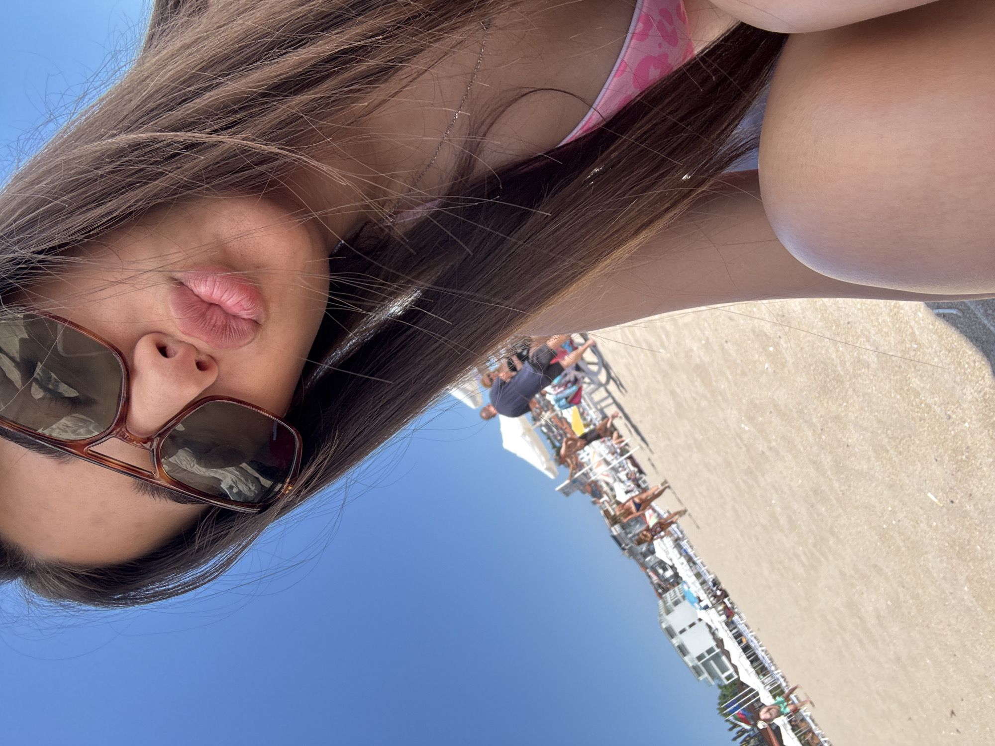 A close-up, high-angle selfie is captured on a sunny day on a beach in Sozopol, Bulgaria. The image focuses on the upper torso of a person wearing sunglasses and a pink bikini top, with their lips puckered. Their long, brown hair frames their face and spills over their shoulder. The background shows a crowded beach with people, umbrellas, lounge chairs, and buildings lining the coastline. The sky is clear and blue.