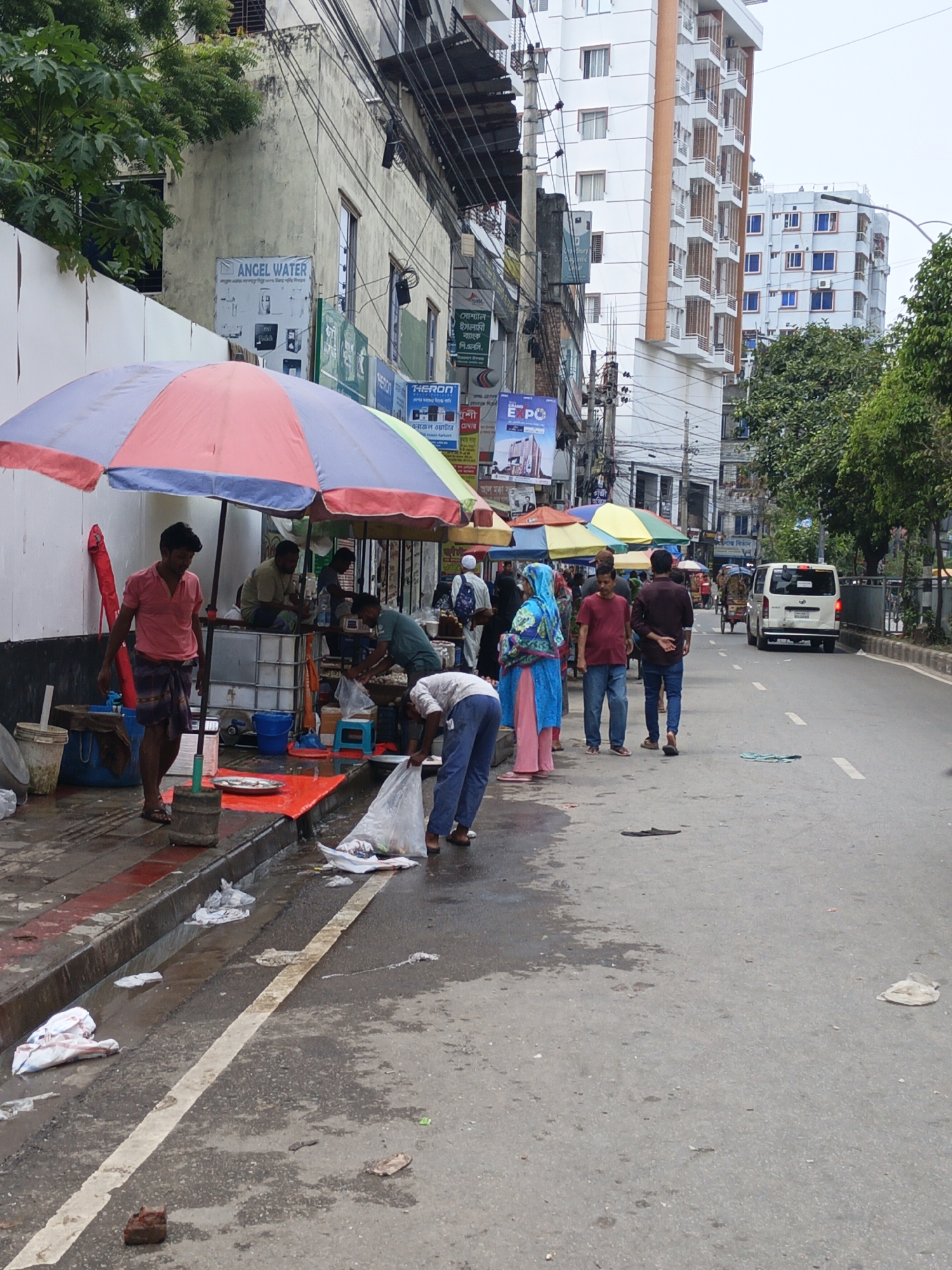 Here's a detailed description of the image:
**Scene Overview:**
The image captures a street scene likely in a South Asian city, possibly Bangladesh. It appears to be a busy commercial area with street vendors selling fish on the sidewalk. A road runs alongside the sidewalk, with vehicles and pedestrians navigating the scene.
**People:**
* Several men are gathered around fish stalls, presumably vendors and customers.
* One man wears a pink shirt and a dark patterned sarong.
* One man is bending over, appearing to be handling a plastic bag.
* A woman in a blue and pink headscarf stands with others walking on the sidewalk.
* People are casually dressed in typical South Asian attire.
**Objects:**
* Fish stalls are set up under colorful umbrellas to provide shade.
* Plastic bins and tanks are visible, likely holding water for keeping the fish fresh.
* Several white plastic bags are scattered near the curb, possibly containing fish or refuse.
* A white van is driving on the road.
* Buildings line the street, including some with signs, such as one for "Angel Water". Other signs in the Bengali script are also present.
* A brick sits on the street near the curb.
**Actions:**
* Vendors are presumably selling fish from their makeshift stalls.
* A man is bending down, likely handling fish he has just purchased.
* People are walking along the sidewalk.
* Vehicles are driving on the road.
**Interactions:**
* Customers are interacting with the fish vendors.
* Pedestrians are walking past the stalls.
* Vehicles are sharing the road with pedestrians and rickshaws.
**Environment:**
* The street is wet, suggesting it may have recently rained or been washed.
* There is trash and debris scattered along the sidewalk and street.
* Buildings of various heights line the street.
* Power lines and utility poles are visible overhead.
* The overall atmosphere suggests a bustling and somewhat chaotic urban environment.