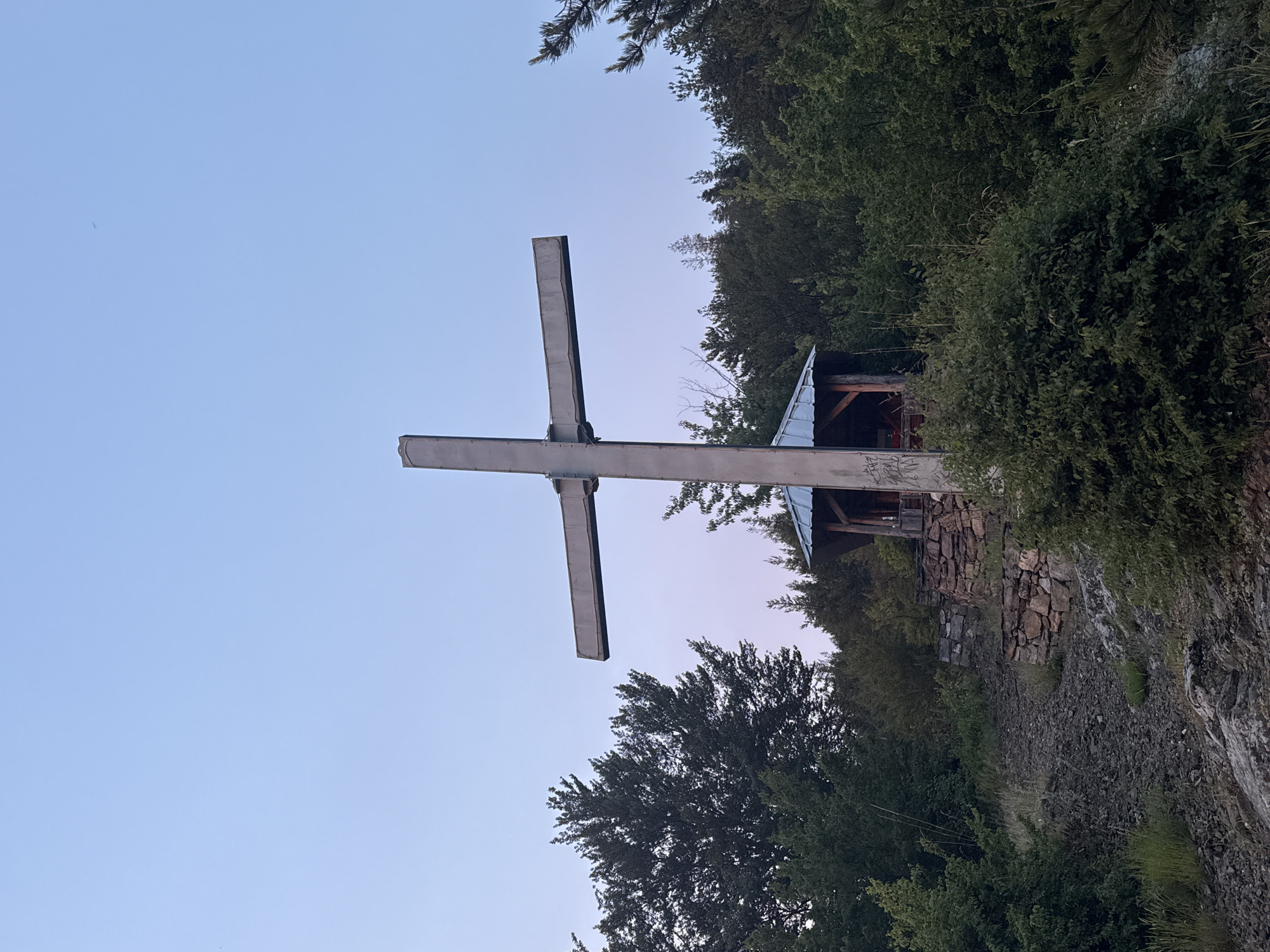 The image depicts a large, metallic cross situated outdoors in a rocky, wooded area. The cross is centrally positioned and appears to be made of concrete or a similar material, with a rough, textured surface. Beneath the cross and slightly to the right, there is a small, open-sided shelter with a pitched roof made of corrugated metal. The shelter has wooden support beams and a floor. The ground leading up to the shelter is covered in loose stones and rocks, with patches of green grass and bushes growing throughout. A stone wall, constructed with irregular stones, is visible behind the path and to the right of the shelter, forming a terraced effect. The background is filled with dense green trees and foliage, characteristic of a forest. The sky above is a clear, pale blue, transitioning to a faint pinkish hue near the horizon, suggesting either dawn or dusk. There are no people visible in the image. The location is identified as Strazha, Bulgaria.