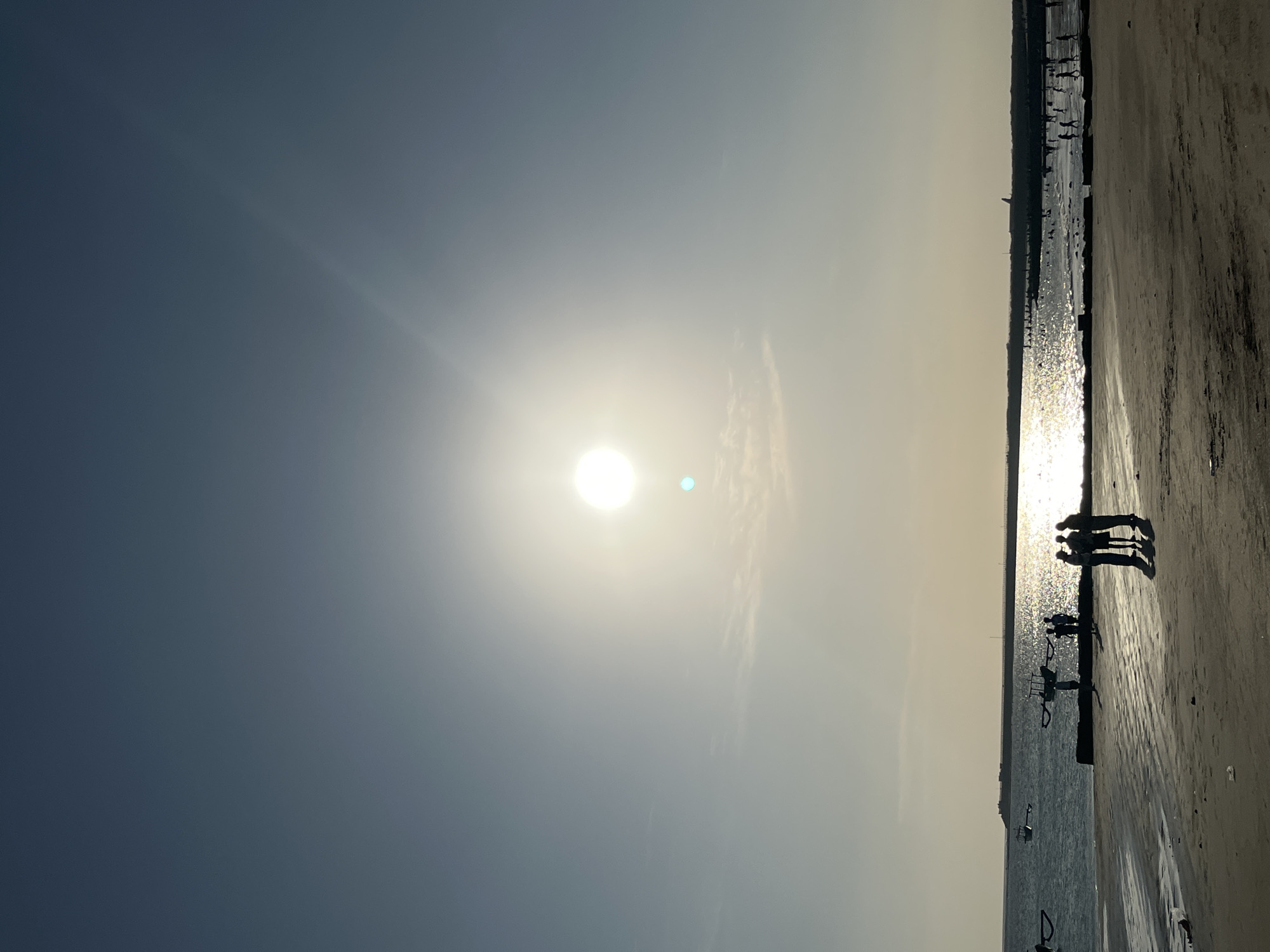This vertical, silhouette-style photograph captures a serene beach scene at sunset in Kuta, Indonesia. The sky dominates the upper half of the image, transitioning from a deep blue at the top to a lighter, hazy blue near the horizon. The sun, a bright, almost white orb, is positioned slightly above the center of the frame, casting a strong glare and a shimmering reflection on the water. A faint sun flare emanates diagonally upwards to the right.
The middle ground features the ocean, with the sun's reflection creating a bright, sparkling path across its surface. Small waves are visible lapping at the shore. Along the water's edge, a line of people are silhouetted against the bright water and sand, some standing individually and others in small groups. In the foreground, a sandy beach is depicted with subtle textures from the wet sand. Several dark, rectangular objects, possibly discarded debris or remnants of structures, are scattered on the sand.
In the far distance, a long, man-made structure, likely a pier or breakwater, stretches across the horizon. Boats with distinct outrigger designs are visible on the water, some closer to the shore and others further out. The overall mood of the image is peaceful and tranquil, emphasizing the natural beauty of the sunset at Kuta beach.