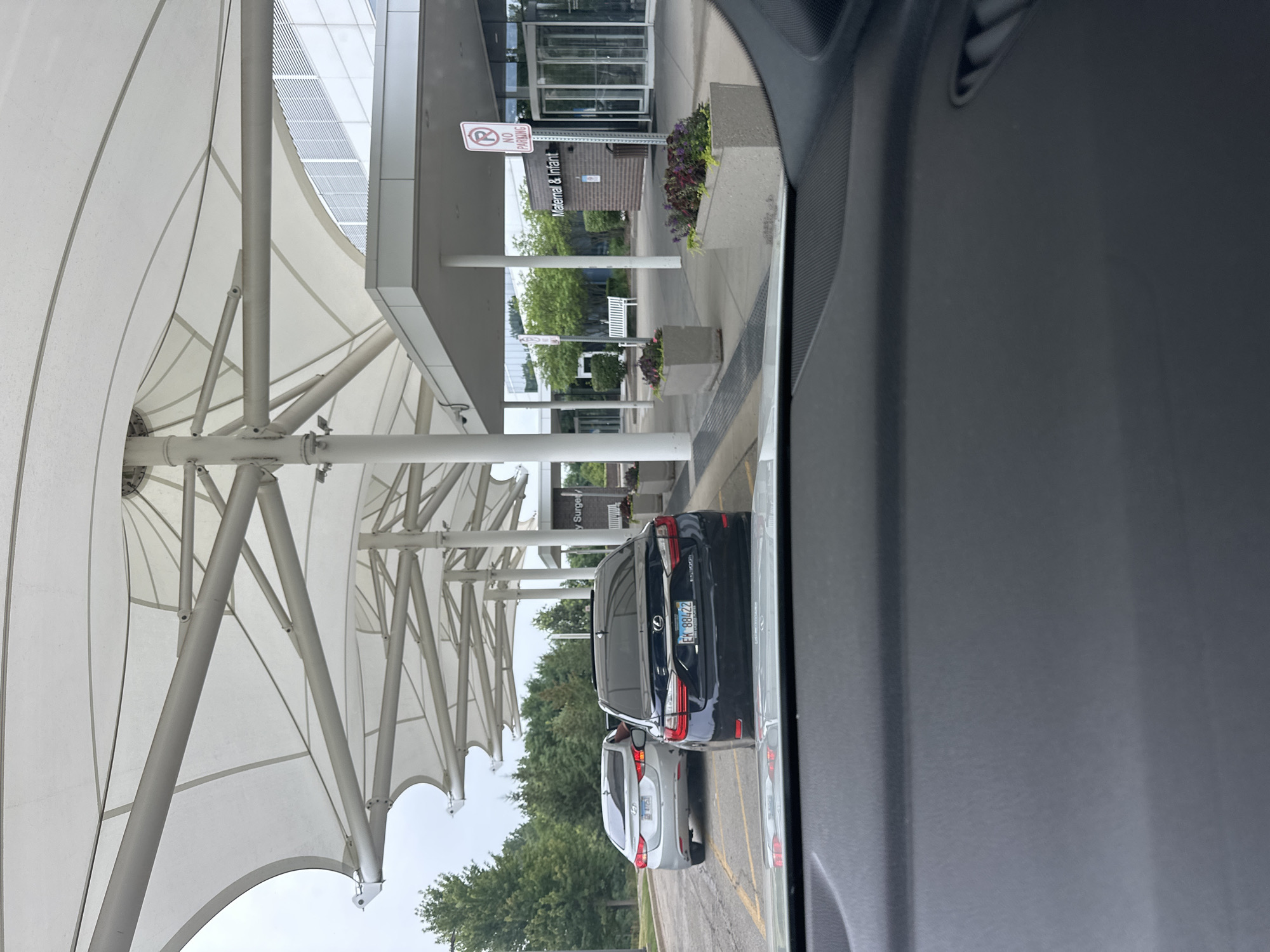 The image depicts the exterior of a hospital building with a large, white, canopy-like structure extending over a drop-off or waiting area. In the foreground, the dashboard of a vehicle is visible. Two cars are parked or waiting in front of the building. The car on the left is silver, and the car on the right is black. The building has a modern architectural design with large glass windows and doors. A sign on the building reads "Maternal & Infant." There are planter boxes with flowers along the walkway. The overall environment appears to be a typical hospital entrance. The location is identified as Hoffman Estates, United States of America.
