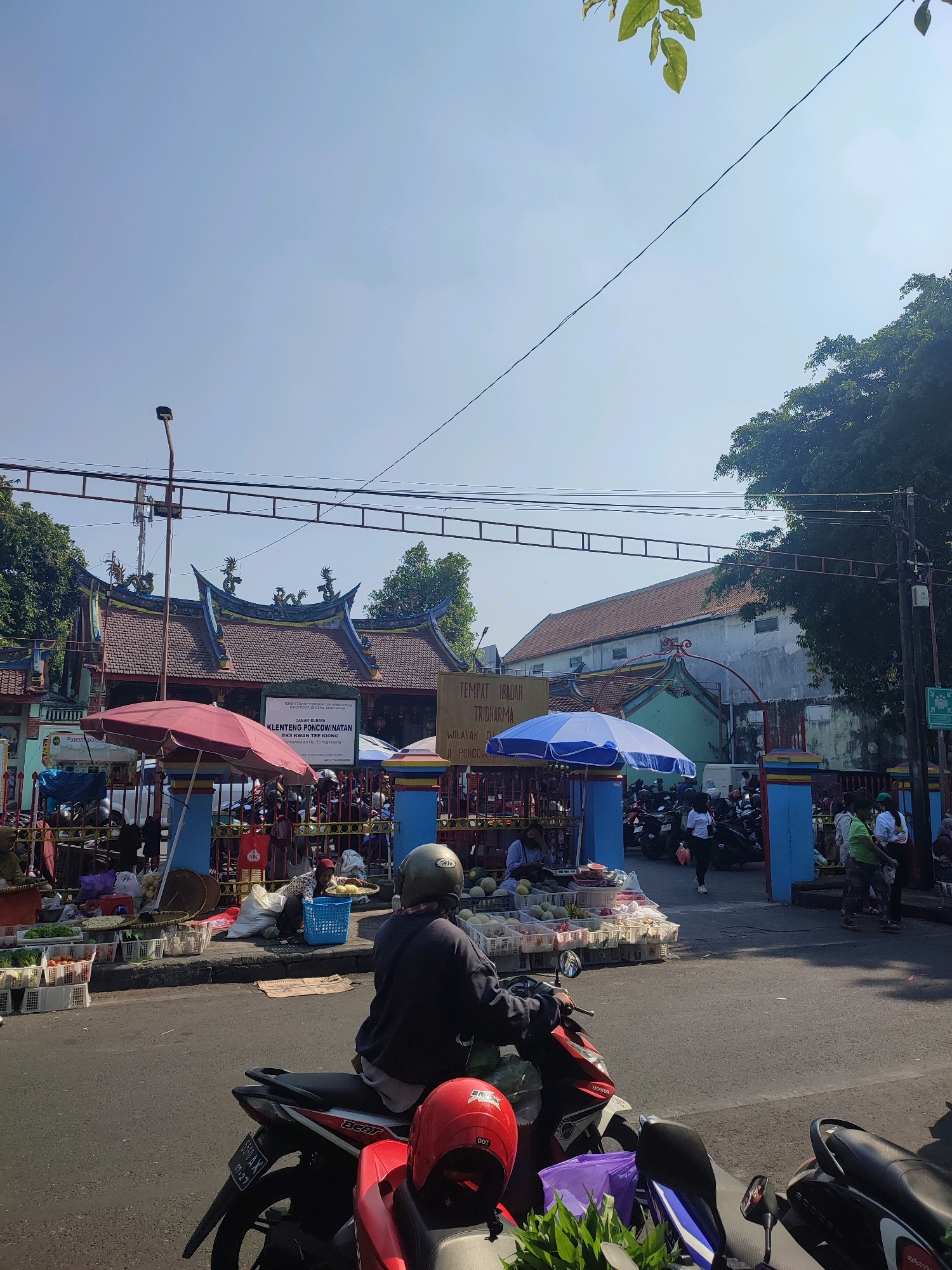 The image captures a street scene in Yogyakarta, Indonesia, featuring a market area in front of a temple. The architecture of the temple, with its decorative roofs and dragon motifs, suggests a Chinese influence, likely indicating it is a Klenteng or Chinese temple. A sign on the temple reads "TEMPAT IBADAH TRIGHARMA" and "KLENTENG PONCOWIHATAN". The market stalls are set up along the roadside, displaying fruits and vegetables in white plastic crates and woven baskets. Two individuals are seated behind the stalls, while others are browsing or walking through the market. Several motorcycles are parked, with one prominently in the foreground, its rider wearing a helmet. The day is bright and sunny, with a clear blue sky. The street is paved, and trees are visible in the background, casting shadows. The overall impression is of a bustling local market in a culturally rich urban setting.