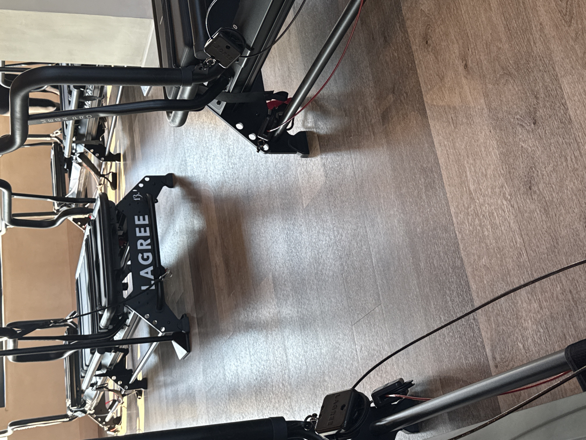 This image depicts the interior of an unoccupied Lagree fitness studio in London, United Kingdom. Several black Lagree Megaformer machines are arranged on a light-to-medium brown wooden plank floor. The branding "LAGREE" is visible in white capital letters on the base of a central machine, and "MEGAFORMER" is readable vertically on an upright handle of a machine to the right. The machines consist of black frames, padded carriages, handles, and associated black and red cabling. Natural light enters from a window on the left, illuminating the scene and creating shadows on the floor and equipment. The walls are light-colored. No individuals are present.