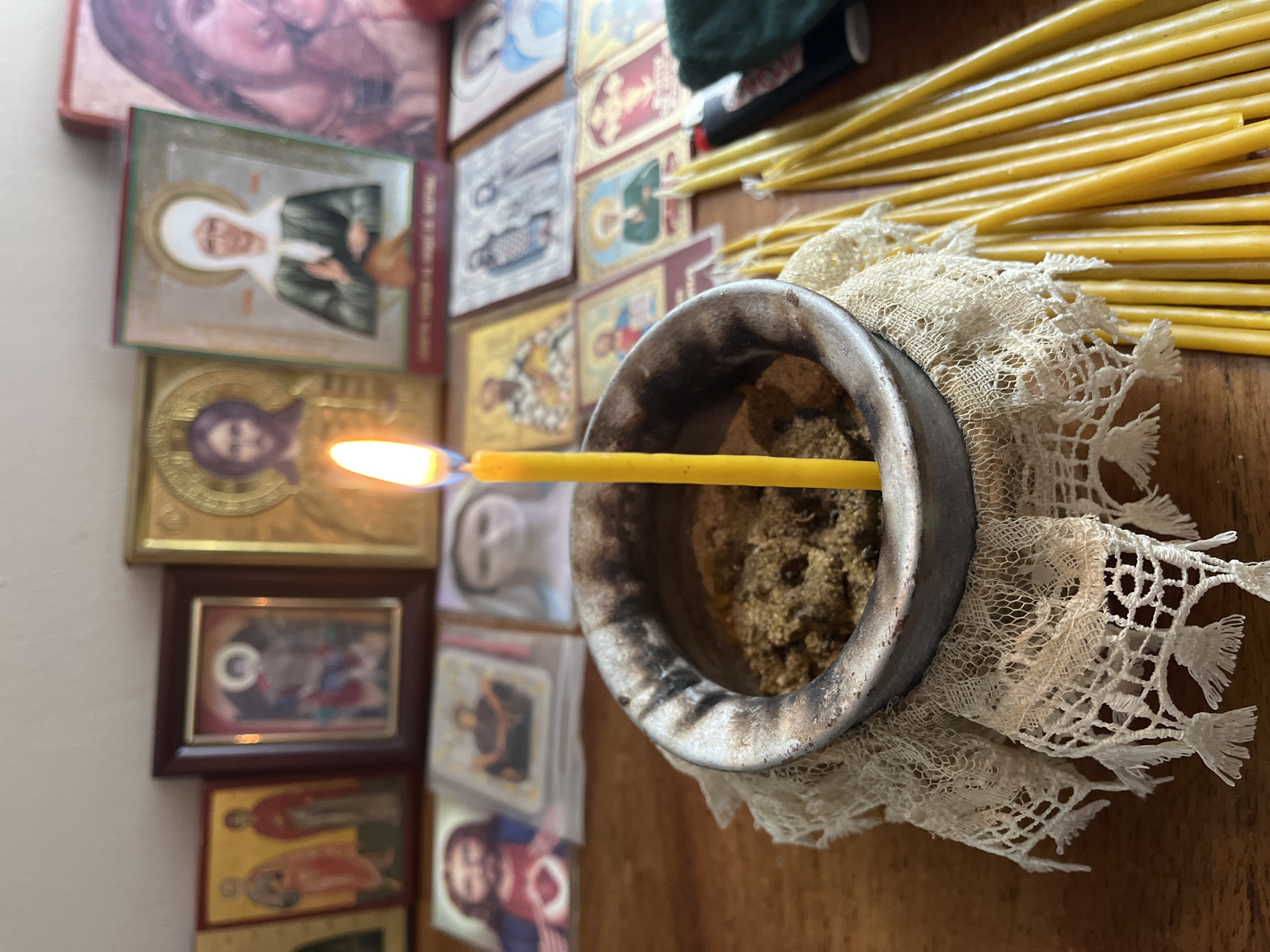 The image depicts an altar or devotional space with several religious icons and a lit candle. In the foreground, a yellow candle burns brightly in a small, tarnished metal censer filled with a granular substance. The censer rests on a piece of white lace with a tasseled edge. To the right of the censer, a bundle of unlit yellow beeswax candles is arranged. In the background, a collection of framed and unframed religious icons, primarily depicting religious figures like Jesus and Mary, are displayed. The icons vary in size and style, some appearing older and more ornate than others. The overall setting suggests a personal or religious space for prayer or contemplation. The location is listed as Unknown, Unknown.