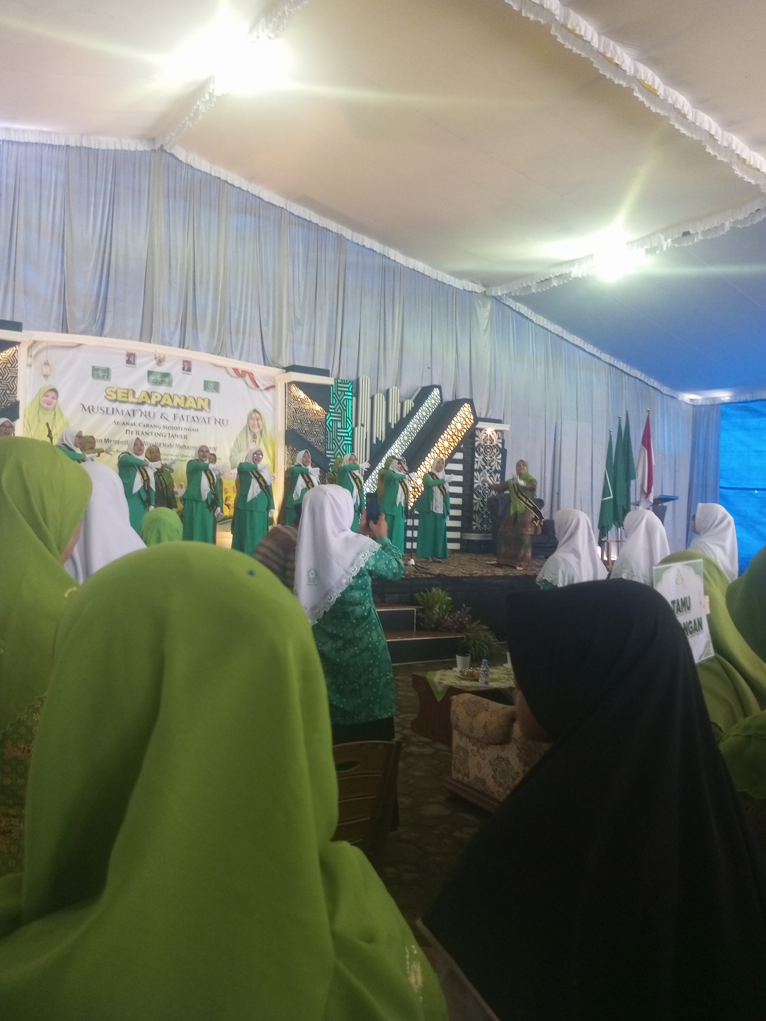 The image depicts an indoor event, likely a ceremony or presentation, held within a tent or covered venue in Klaseman, Indonesia. A stage is set up at the back, adorned with banners and decorative elements. Several individuals, primarily women wearing hijabs and green attire, are gathered in front of the stage. Some are seated, while others appear to be part of a performance or presentation on stage. The backdrop of the stage features a large banner with text, indicating the event name as "SELAPANAN MUSLIMAT NU & FATAYAT NU DI RANTING JAWAR." Several flags are also visible on the stage, some of which are green. The environment appears to be a gathering of a religious or community organization, possibly related to Nahdlatul Ulama (NU) and its affiliated women's organizations. The lighting suggests an indoor setting, potentially a community hall or an open-air venue covered by a tent. The overall atmosphere seems formal and organized, with attendees dressed in traditional or formal clothing.