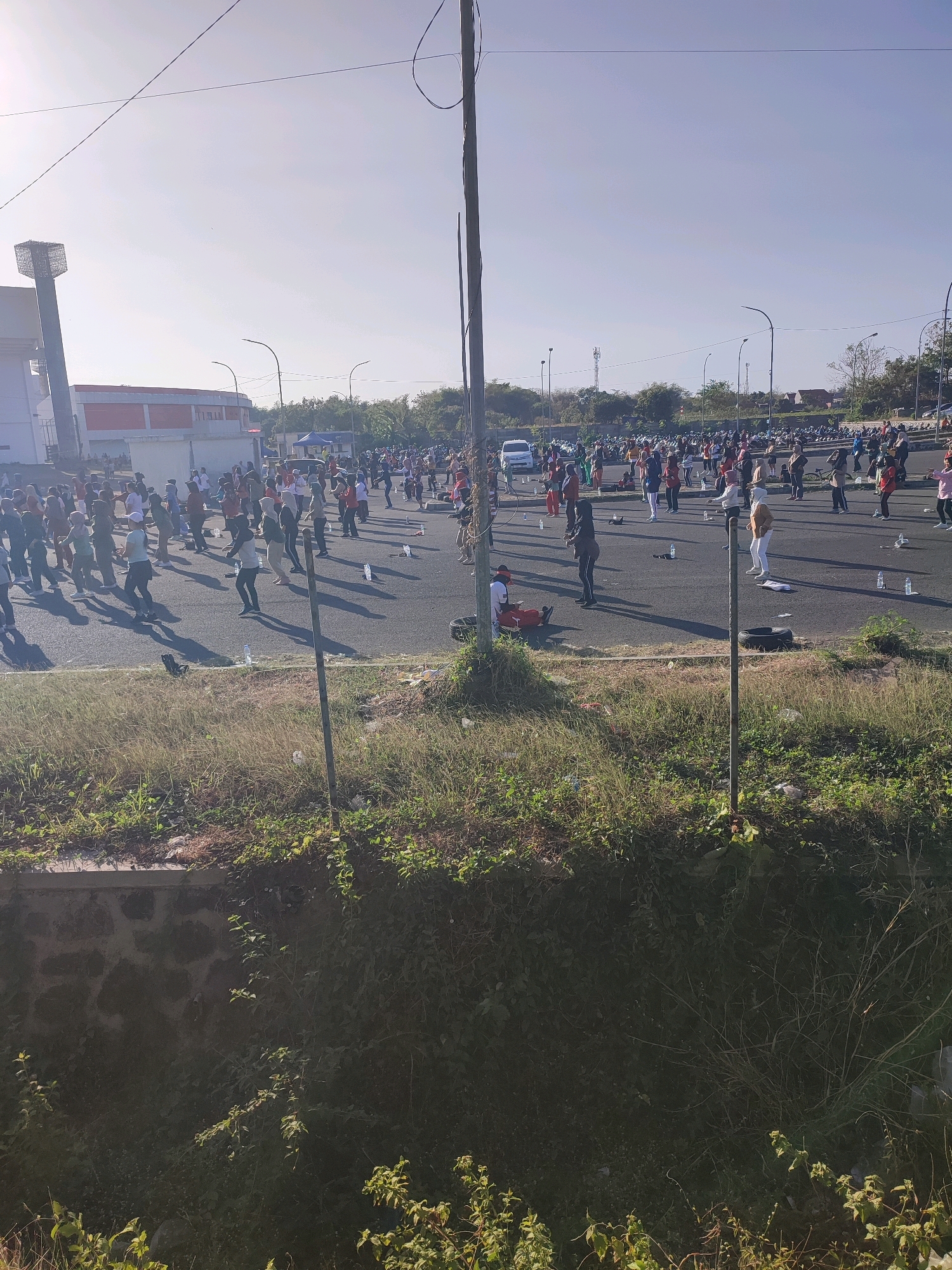 The image depicts a large outdoor gathering of people, likely participating in a group exercise activity. The scene is set in a paved outdoor area, possibly a parking lot or street, in Watubelah, Indonesia. The ground is asphalt, with some grass and vegetation visible in the foreground and along the edges. A wall made of stone and overgrown with plants is present in the lower part of the image. In the background, several buildings are visible, including one with a distinctive tower structure and another with a red roof. Streetlights line the area where the people are gathered, and several parked vehicles, including cars and motorcycles, are scattered throughout the scene. The sky is clear and bright, suggesting it is daytime, likely morning due to the angle of the sunlight casting long shadows.
The people are spread across the paved area, mostly in a state of physical activity. Many are standing in rows, facing in the same direction, and appear to be performing synchronized movements. Some individuals are wearing athletic or casual clothing, with a variety of colors and styles. There are water bottles scattered on the ground among the participants. A few individuals are seated or in more relaxed poses. The overall impression is one of organized community activity. The presence of a tall pole with wires running across the sky, and a few smaller poles, suggests an urban or semi-urban environment.