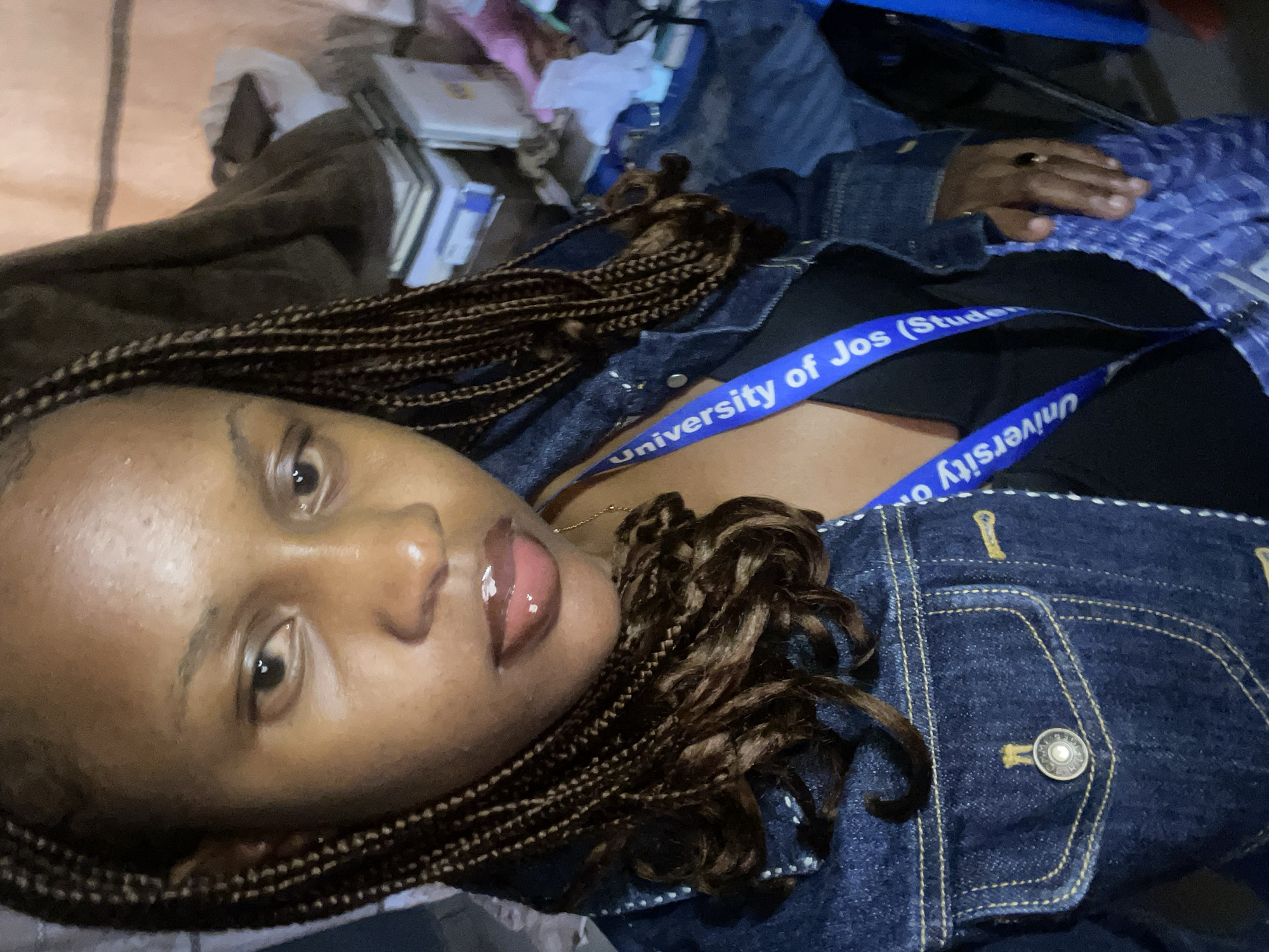 The image shows a close-up of a young woman with braided hair wearing a denim jacket. She is wearing a black top underneath the jacket and a blue lanyard with "University of Jos (Student)" written on it. She is wearing a ring on her right hand. The background is cluttered with books, blankets, and other items, suggesting an indoor setting. The lighting is dim. The location is identified as Rafin Par, Nigeria.