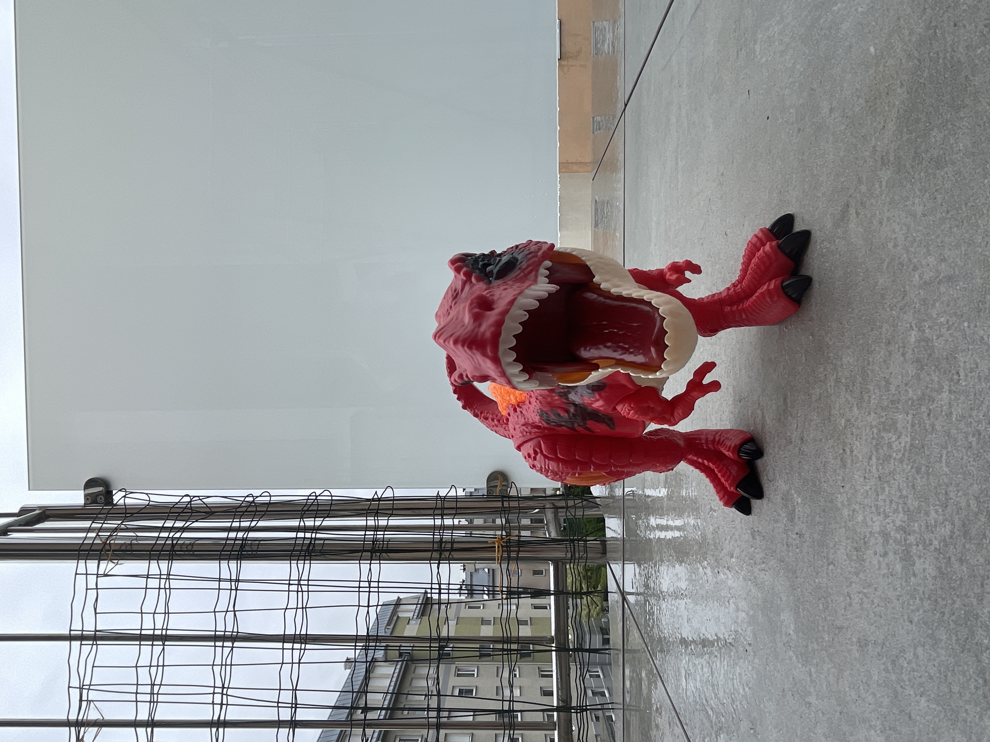 An outdoor, eye-level, medium shot captures a bright red, plastic dinosaur toy in the foreground, positioned centrally on a wet, gray, tiled surface. The dinosaur, with its mouth wide open, revealing white teeth and a red interior, is the sole subject. Its left leg is slightly bent, and its right leg is also bent, with both feet firmly planted on the ground. The dinosaur has black claws and a patterned detail on its back, as well as orange accents near its neck.
Behind the toy, a thin, black metal fence runs vertically and horizontally, creating a grid-like pattern. Beyond the fence, residential buildings with multiple windows are visible, suggesting an urban setting. The sky is overcast and gray. The overall environment appears to be a balcony or a rooftop patio, given the presence of the railing and the elevated perspective. The wet surface indicates recent rain or water.