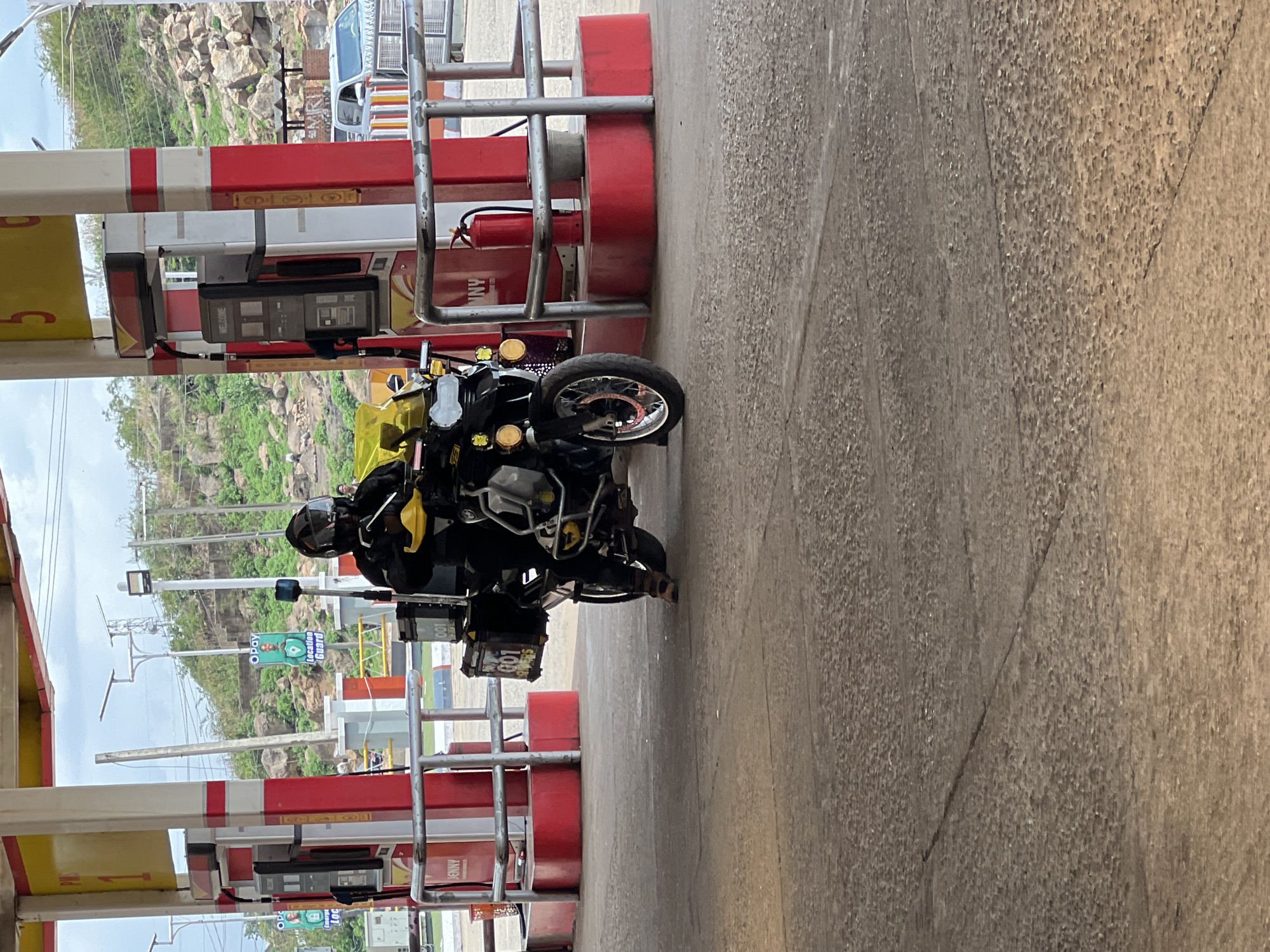 The image shows a motorcyclist at a gas station in Gura Top, Nigeria. The motorcycle, a black and silver touring model with saddlebags, is parked next to a red and white gas pump. The rider, wearing a black helmet and gear, is seated on the motorcycle. The gas station is identified by the "Jenny" logo on the pump. In the background, there's a hillside with trees and power lines. Other vehicles are partially visible in the distance. The ground is concrete. A sign for "Opay Location Guard" is visible on a pole in the background.