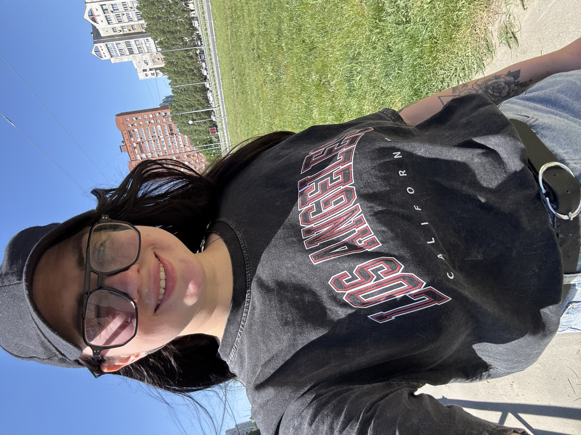 A woman is taking a selfie outdoors. She is wearing glasses and a baseball cap. She is also wearing a black t-shirt with "LOS ANGELES CALIFORNIA" printed on it. The background features apartment buildings and a grassy area. The city and location are unknown.