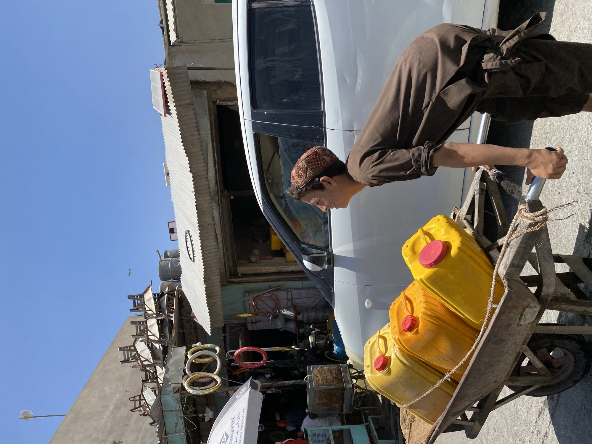 The image captures a street scene in Kandahar, Afghanistan, under a bright blue sky. In the foreground, a young man wearing a brown tunic and a traditional kufi cap leans over a rustic wooden cart. He is actively engaged in pushing or maneuvering the cart, which carries three large, yellow jerrycans secured with rope. The jerrycans have red caps and appear to be filled with a liquid. Behind the cart and the young man is a white vehicle, partially visible. The background reveals a bustling urban environment with buildings characterized by corrugated metal roofs and older structures. Some of these buildings house shops, with goods like coiled wires and what appear to be chairs stacked on top of one of the structures. The overall impression is one of everyday life and commerce in a city.