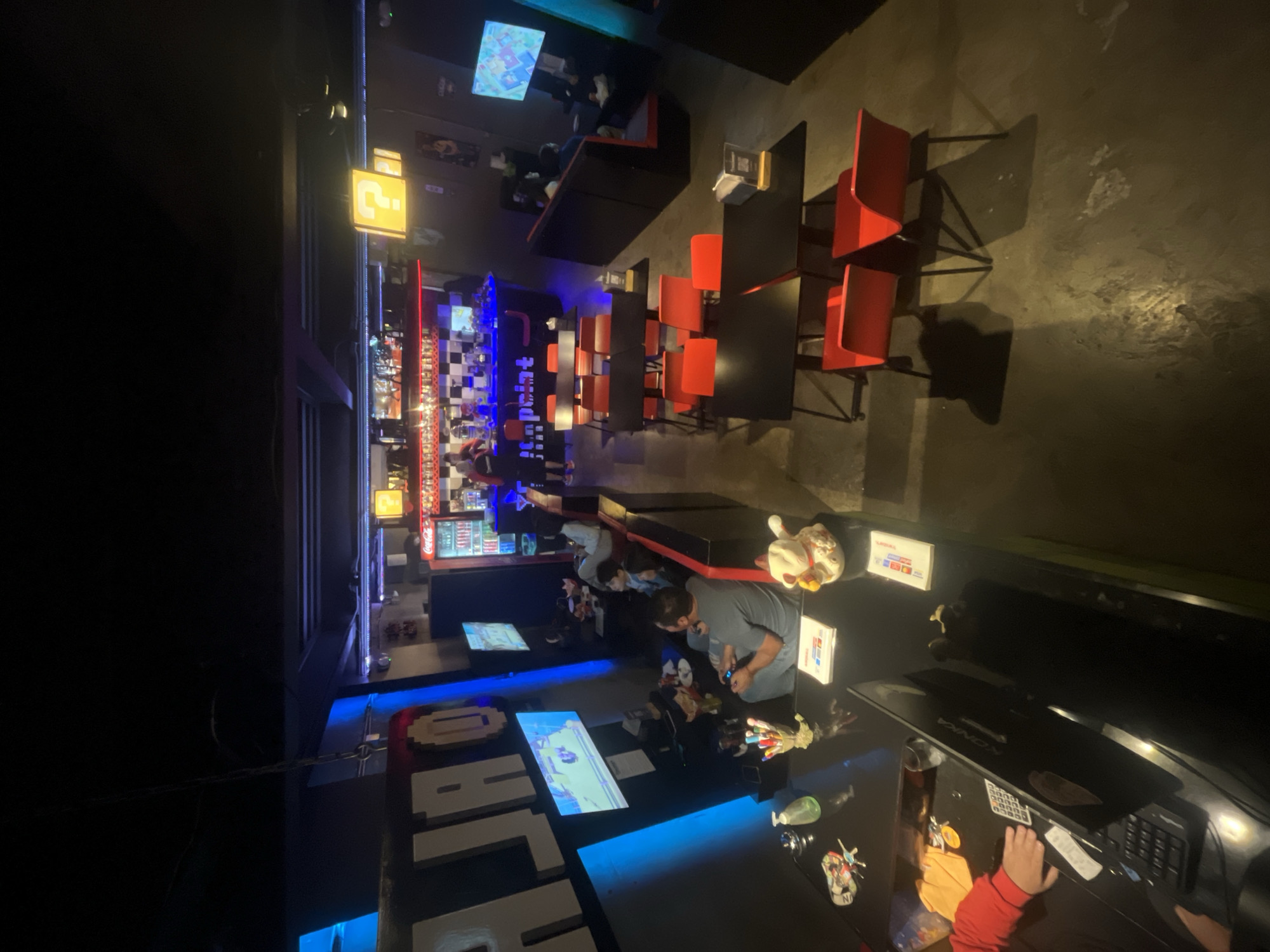 The image depicts the interior of a dimly lit entertainment venue that appears to be a video game cafe or bar, possibly located in Iquique, Chile. The scene is viewed from an elevated perspective. In the foreground, a black table is visible with several items on it, including pens, a bottle of hand sanitizer, keys, and what looks like a calculator and a laptop. A person in a red long-sleeved shirt is partially visible on the left, with their hands near the calculator and laptop. To the right of the table, a plush toy resembling a cartoon character sits next to some papers.
Further back, several black tables are arranged with bright orange chairs. A bar area with a checkered pattern on its counter is present in the mid-ground, with patrons and staff visible. Behind the bar, a Coca-Cola branded refrigerator is stocked with drinks. Yellow signs shaped like question marks, reminiscent of the Super Mario video game series, are mounted on the wall. Multiple televisions are strategically placed throughout the venue, some displaying video games, indicating the primary purpose of the establishment. In the background, a dark seating area with red accents is occupied by individuals, some of whom appear to be playing video games. The overall ambiance is casual and designed for entertainment.
