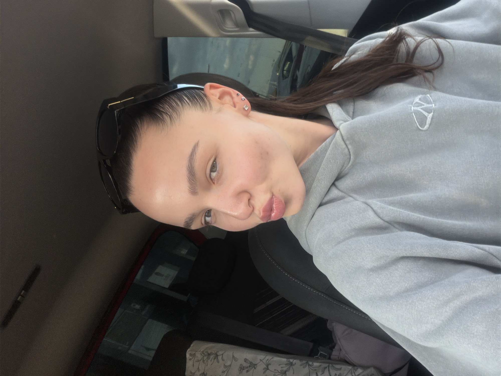 A young woman is captured in a close-up selfie taken inside a car. She is posing with a "pouty" expression, her lips slightly pursed. Her dark hair is slicked back, with sunglasses perched on top of her head. She has multiple small, clear, and one dark stud earring in her left ear. She wears a light grey hooded sweatshirt featuring a distinct white embroidered logo on the chest, which appears to be a stylized letter or abstract symbol.
The setting is the interior of a vehicle, suggested by the visible car seat, door panel, and roof lining. Through the car window in the background, blurred outdoor elements are visible, indicating it is daytime. A smartphone with a red case is also partially visible on the left side of the frame. The woman's skin shows a natural, slightly dewy appearance. The photo is rotated, presenting the woman's head tilted to the right. The overall location is Sofia, Bulgaria.