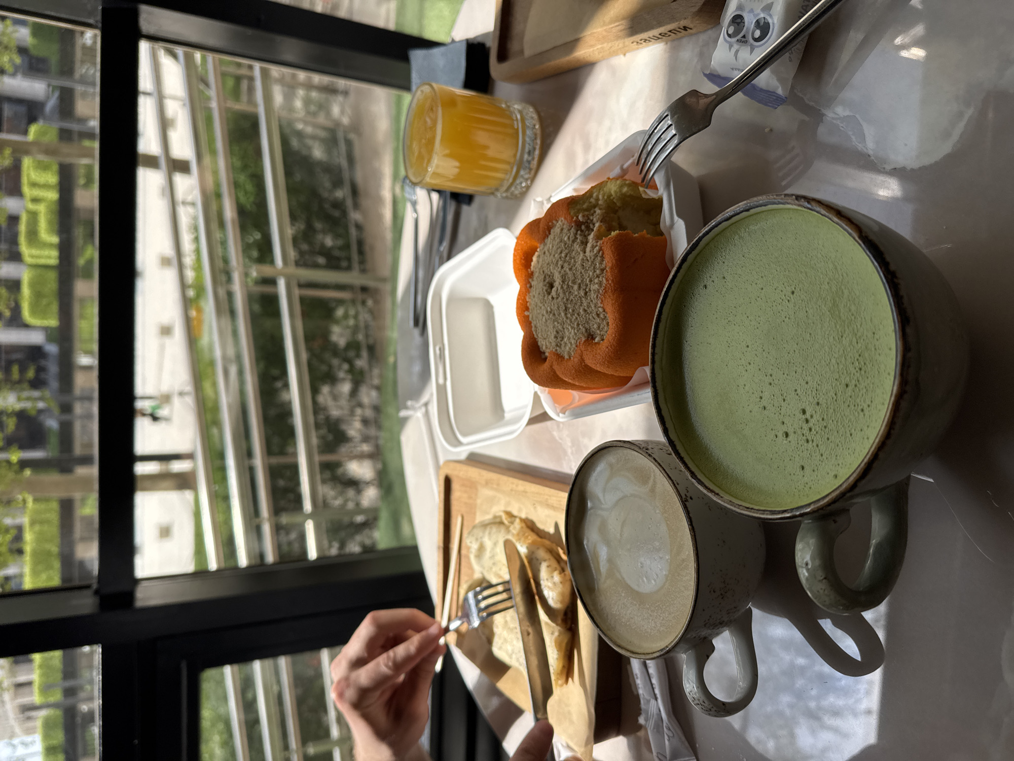 The image displays an indoor dining scene, likely at a cafe or restaurant, bathed in natural sunlight. A person's hands are visible in the lower left, holding a fork and knife, poised to eat a meal.
On the table, an array of food and beverages is arranged. In the foreground, a light-colored, foamy drink, possibly a latte or cappuccino, sits in a mug. Next to it, a second mug holds a vibrant green, frothy beverage, characteristic of a matcha latte. Behind these, a white package adorned with cartoon eyes and the word "OPEN" is visible, alongside a metal fork.
A small glass of orange juice is placed further back on the left. In the center, an orange-colored pastry, resembling a muffin or small cake with a crumbly topping, rests in a white disposable container. Another empty white container is also on the table. The person's cutlery is engaged with what appears to be folded crepes or pancakes, served on a wooden board. The board features text engraved in a non-Latin script, which resembles the Cyrillic word "зацепи".
Through the large, dark-framed window, an outdoor scene unfolds, showcasing tall buildings and abundant green trees and foliage, suggesting an urban or suburban setting. The bright, direct sunlight streaming into the room indicates it is daytime, possibly morning or early afternoon. The scene conveys a relaxed meal in a well-lit environment.