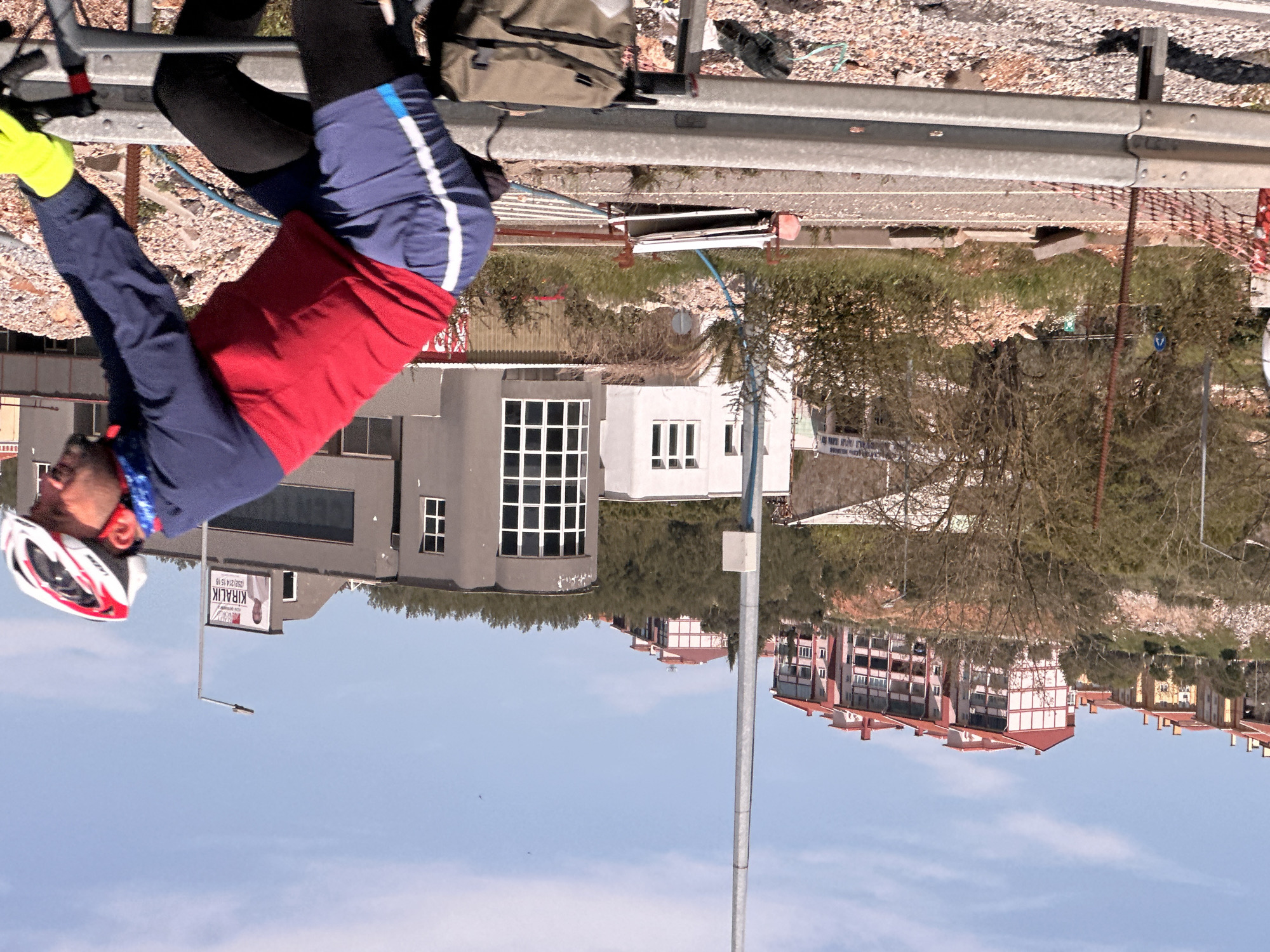 A person wearing a red, white, and blue cycling outfit and a helmet is sitting on a metal railing in Menteşe, Turkey. The person is wearing bright yellow gloves and is resting their arm on the rail. Behind the person, there is a paved road with gravel on the side and green vegetation. There are several buildings in the background, including a gray building with a sign that reads "KIRALIK." Beyond the buildings, there are trees and houses on a hillside. The sky is light blue with a few clouds.