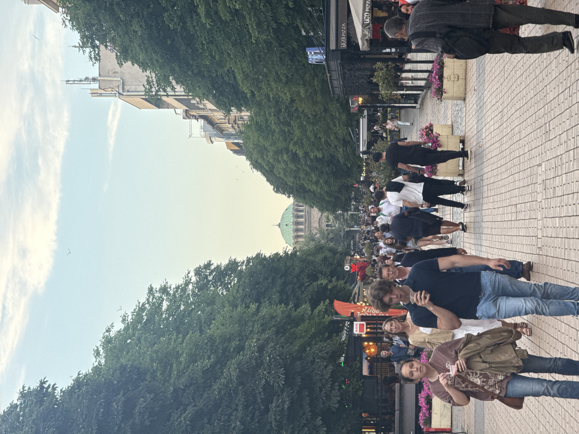 In Sofia, Bulgaria, on Vitosha Boulevard, a pedestrian street lined with trees and shops, a crowd of people is walking. In the background, the dome of Alexander Nevsky Cathedral is visible. The image captures a lively atmosphere with people enjoying their evening stroll. The street is paved with cobblestones, and there are planters filled with pink flowers along the sides. Several businesses, including restaurants and cafes, are open and have outdoor seating. The sky is a soft blue with scattered clouds.