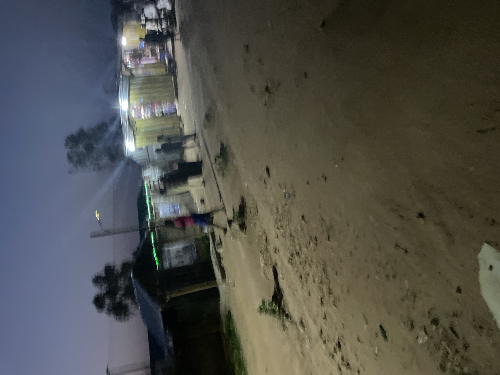 The image depicts a dimly lit street scene at dusk or dawn in an urban or semi-urban environment. The ground is composed of dirt and gravel, with scattered debris. On the right side of the street, there are several wooden market stalls or shops, illuminated by artificial lights, displaying various goods. Smoke or steam is visible rising from one of the structures. Several blurred figures of people are present, some appearing to be walking or standing near the stalls. On the left side of the street, there is a dark, low-lying structure, possibly a building or another stall, with some green lights visible. A lamppost with a lit bulb is situated between the stalls and the structure on the left. The sky is a muted purple and grey, suggesting the transition between day and night. The overall impression is of a bustling, albeit somewhat rough, commercial area.