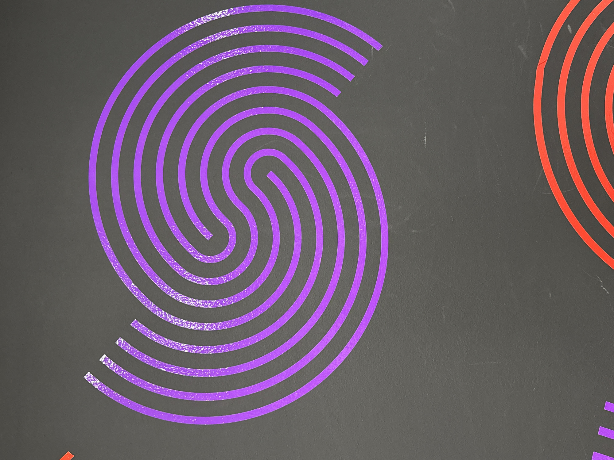 The image shows a close-up of an abstract design on a gray surface. The design consists of concentric lines in purple forming a spiral or maze-like pattern. Part of a similar design in red is visible on the right edge of the image. There are no people, actions, or interactions visible in the image. The location in Chicago is unknown.