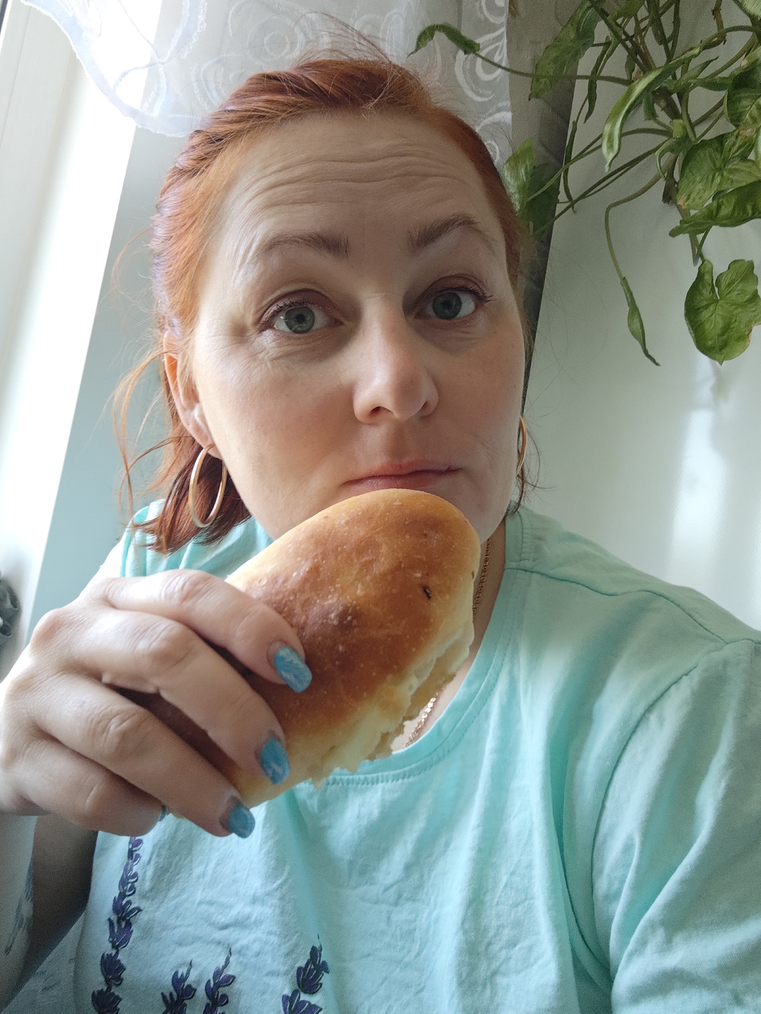A woman with red hair is holding a baked pastry in her left hand, positioning it near her mouth. She is wearing a light blue t-shirt with a floral design, and gold hoop earrings. Her fingernails are painted blue. The background includes a window with a white sheer curtain and a potted plant with green leaves. The location, Фоки, Russia, cannot be determined from the image.